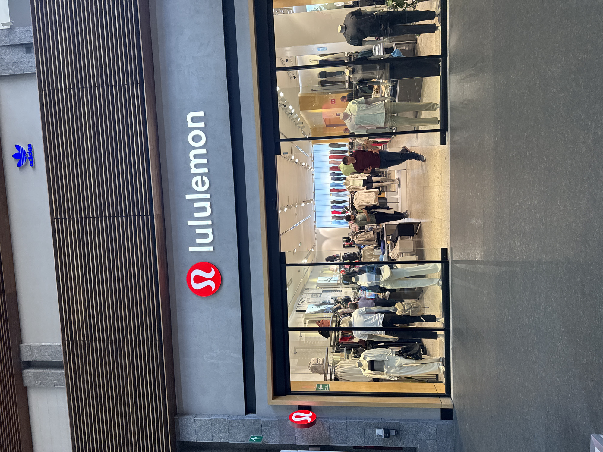 The image shows the exterior of a Lululemon store. The store's name and logo are prominently displayed vertically on the facade. An Adidas logo is placed above the Lululemon store. Inside, mannequins are visible, dressed in various athletic and casual wear. People are walking inside the store. The store appears to be located in a shopping mall or commercial area, with a modern aesthetic, evidenced by the gray concrete flooring. The Lululemon store is located in the Angelópolis Lifestyle Center in Puebla, Mexico.