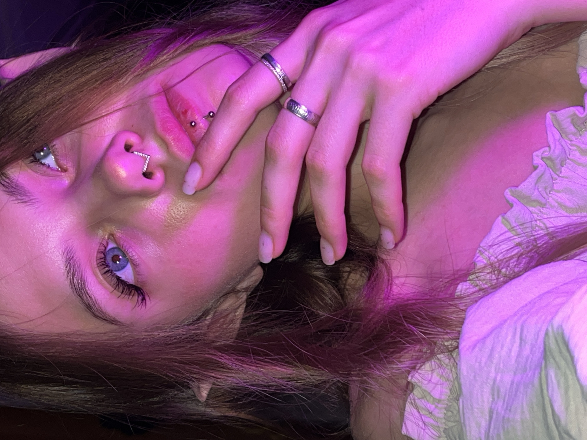 A person is lying down, face partially obscured by a hand adorned with two rings. The face is illuminated with a pinkish-purple light, revealing blue eyes and multiple facial piercings on the nose and lip. Brown hair is visible around the face and neck, and a white patterned garment is partially visible. The environment is dark and undefined. The location Фоки, Russia is not discernible from this image.