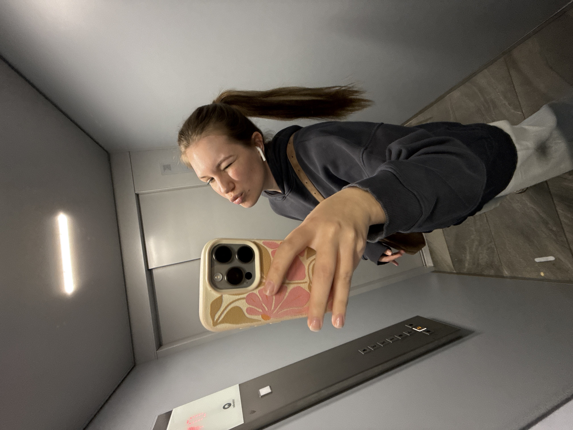 A young woman takes a selfie in what appears to be an elevator. She has brown hair pulled back in a ponytail, wears a dark grey sweatshirt, and has white earbuds in her ear. She is holding a smartphone with a floral-patterned case in her hand, facing towards the elevator panel with buttons. The elevator has grey walls and a wood-patterned floor. The light in the elevator is visible as a vertical streak on the wall. The location appears to be an interior space, likely within a building but there is no specific location data. Therefore I am unable to confirm that it is in Vozdovac Urban Municipality, Serbia.