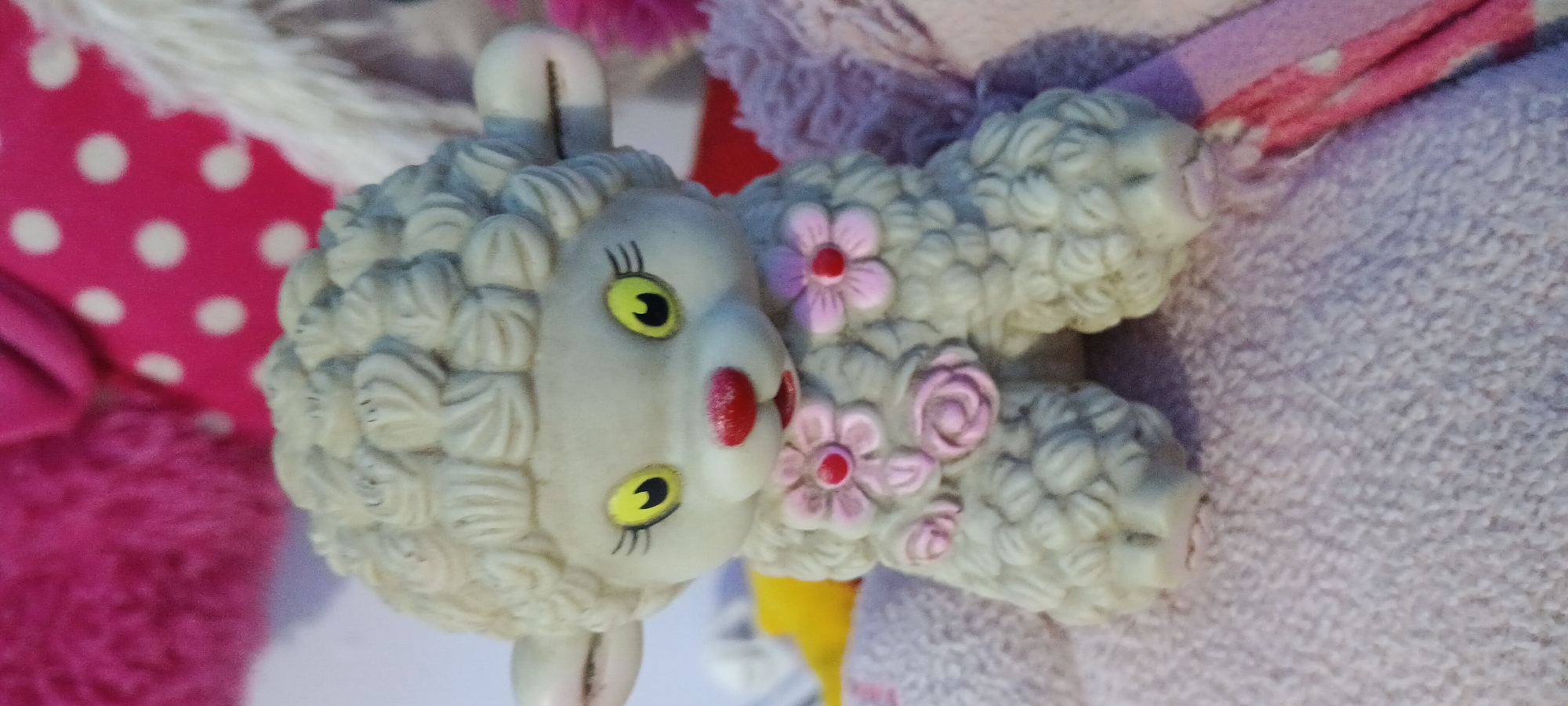 This media file features a close-up, slightly tilted shot of a collection of plush toys. The primary subject is a stylized white lamb toy with a textured, curly wool appearance. It has large, yellow, almond-shaped eyes with black pupils and painted black eyelashes, and a small pink nose with red dots on either side, resembling rosy cheeks. The lamb is adorned with pink flowers with red centers, and what appear to be smaller pink buds or roses on its body.
The lamb toy is positioned amidst other plush toys and fabrics. To the left, a bright pink fabric with white polka dots is visible, along with a darker pink fluffy material. Behind and to the right of the lamb, a predominantly pink and purple plush fabric can be seen, with some striped detailing in pink and possibly white or light pink. The overall setting appears to be indoors, likely a child's room or a crafting space, given the presence of soft toys and fabrics.
The lighting suggests it is daytime, possibly with natural light, as there are soft shadows. There is no discernible activity taking place, as the toys are stationary. No text is visible in the image. The colors are vibrant, with a strong emphasis on pinks and purples, and the bright yellow of the lamb's eyes. The focus is on the texture and detail of the toys. The image is cropped to focus tightly on the lamb and its immediate surroundings.