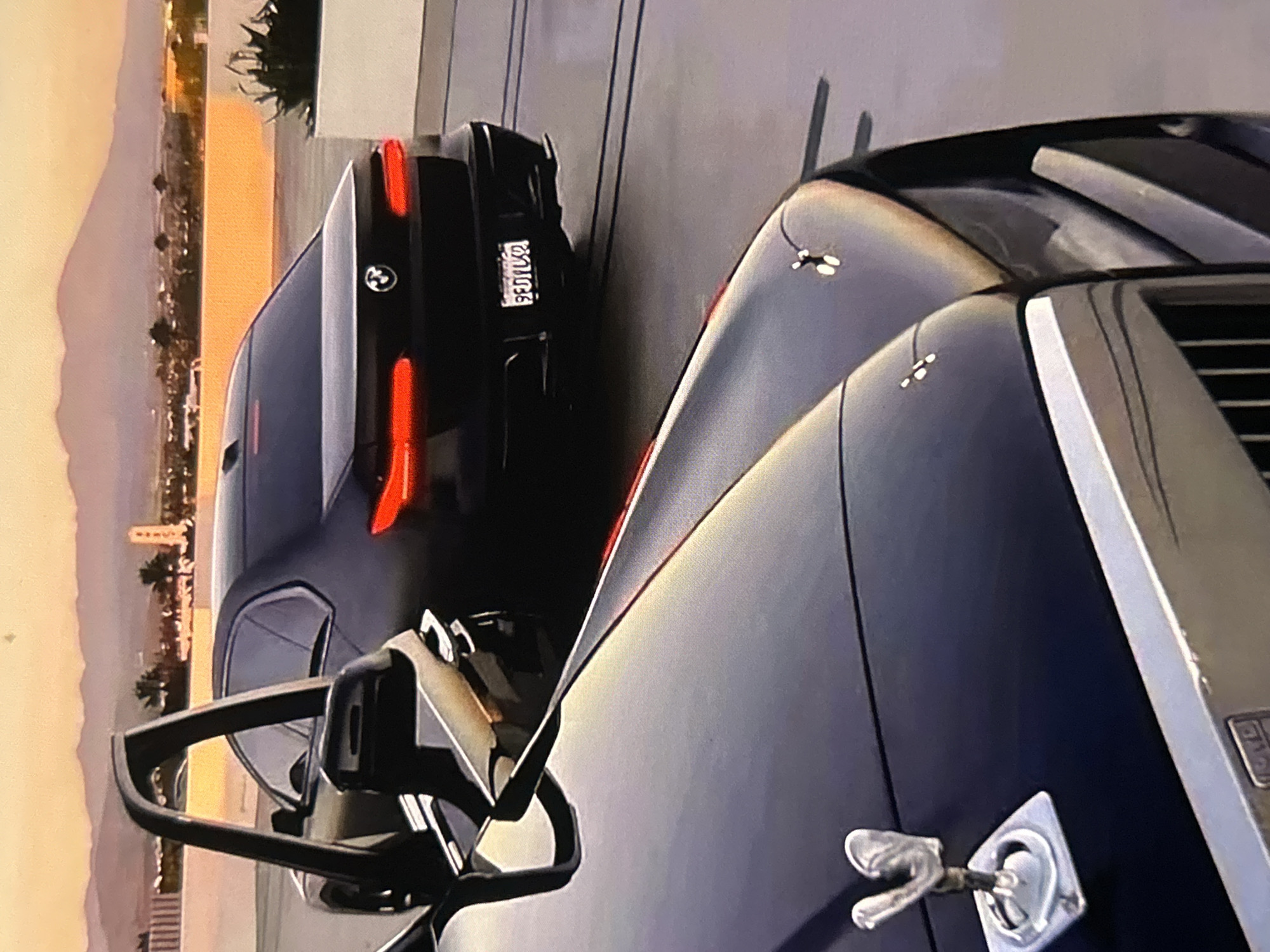 The image displays a scene featuring two luxury cars parked outdoors during either sunset or sunrise, indicated by the warm orange and yellow light illuminating the sky and distant landscape. The visual characteristics suggest this is a still from a video game, noted by the rendering style.
In the foreground, on the right side of the frame, is the front portion of a dark blue or black Rolls-Royce. Its distinctive chrome grille and the iconic "Spirit of Ecstasy" hood ornament are clearly visible, emerging from the car's hood.
Behind and to the left of the Rolls-Royce is a black BMW sedan, viewed from its rear-quarter perspective. The car features prominent red LED taillights and a subtle spoiler integrated into its trunk lid. The BMW emblem is visible on the trunk. The license plate affixed to the rear of the BMW reads "S3IL05".
The cars appear to be stationary, parked on a paved surface next to what might be a building or wall, partially seen on the left. In the background, a cityscape or town with scattered lights is silhouetted against the bright sky, with mountains visible in the far distance. The sky is clear, suggesting fair weather.
There are no specific visual elements within the image that definitively confirm the location as Bania, Bulgaria.