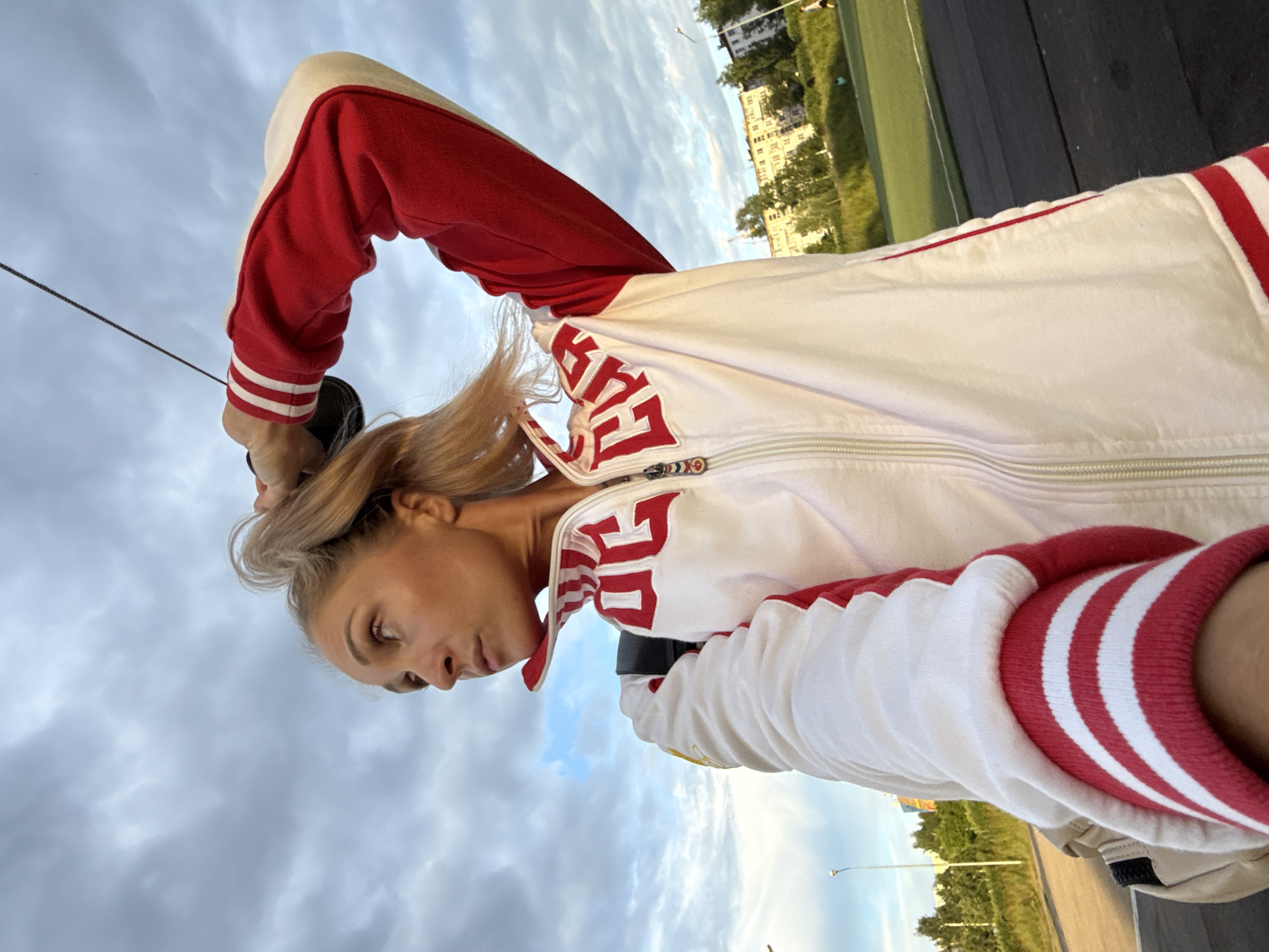 A woman with blonde hair pulled into a ponytail, wearing a red and white athletic jacket, stands outdoors. She is looking to her left, with her right arm raised, touching her hair. The jacket has red trim and a red stripe on the sleeves. A dark backpack is visible over her left shoulder. The background includes a cloudy sky, some trees, buildings, a street lamp, and a green sports field. This image was captured near the Chaikovsky Stadium in Chaikovsky, Russia.