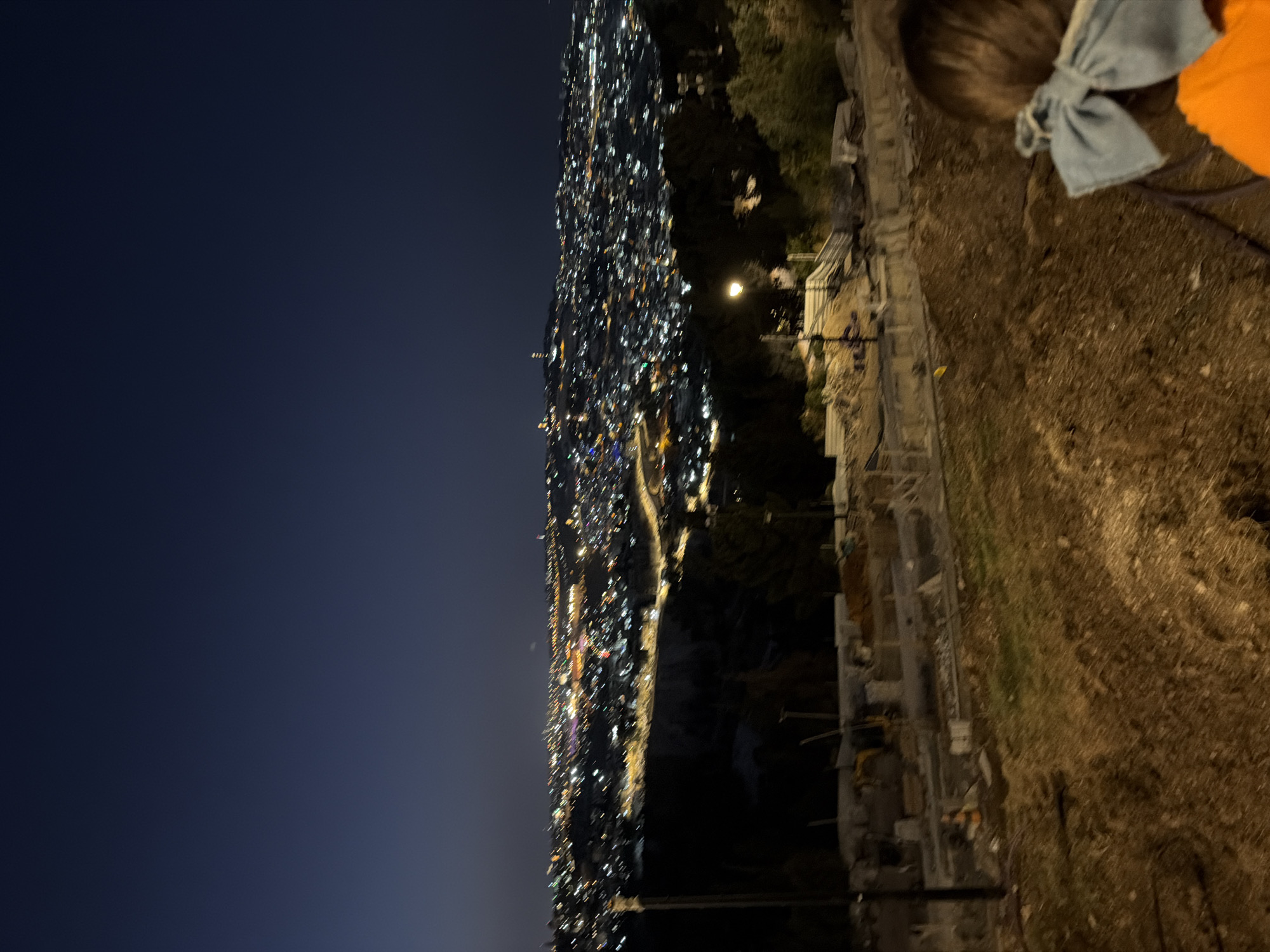 The image captures a panoramic nighttime view of Jerusalem, Israel, from an elevated position overlooking a sprawling cityscape illuminated by countless lights. In the foreground, a partially constructed area is visible, with mounds of dirt, building materials, and construction equipment. A single streetlamp illuminates a small section of this foreground. In the far distance, the lights of the city stretch across the horizon, with notable clusters of lights indicating significant urban areas. The sky is dark, indicating it is nighttime. On the right side of the frame, the back of a child's head is visible, adorned with a light blue bow. The child is wearing an orange shirt. The overall impression is of a vast urban landscape at night, seen from a vantage point with ongoing construction in the immediate vicinity.