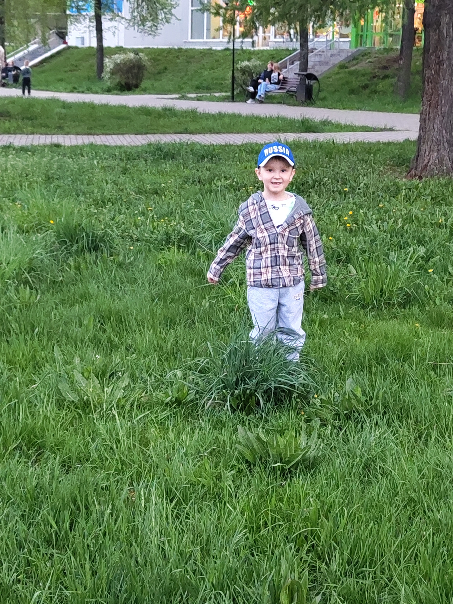 The image shows a child standing in a grassy area, likely a park or green space in Chaikovsky, Russia, indicated by the 'Russia' text on his cap. The child is wearing a blue cap, a plaid shirt over a t-shirt, and gray sweatpants. He is smiling and appears to be facing the camera. The grass is relatively tall and interspersed with some weeds. In the background, there is a paved pathway, a grassy hill, and some trees. Further in the distance, there is a building, and some people are seated on a bench. There's also a slide visible on the left side of the scene. A large tree is on the right side of the picture. The lighting suggests it is daytime.