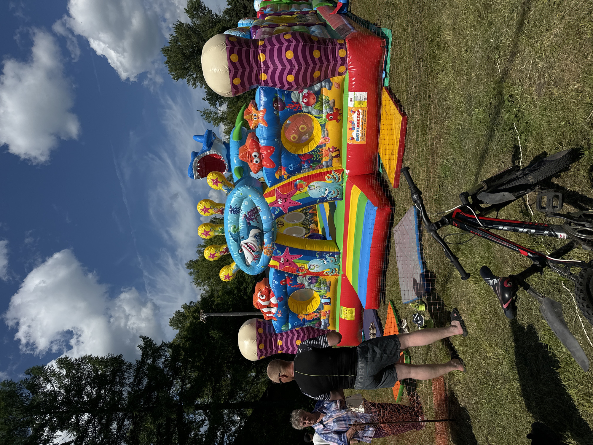 An outdoor shot captures a brightly colored inflatable bouncy castle with an ocean theme, featuring sharks and various sea creatures, set up on a grassy area under a partly cloudy sky. In the foreground, a black bicycle with red accents is parked on the grass, partially enclosed by a safety net. To the left of the bouncy castle, two individuals are visible: an elderly woman in a plaid shirt and a man in a black t-shirt and shorts. The environment appears to be a park or recreational area in the city of Ponazyrevo, Russia, with trees forming a backdrop.