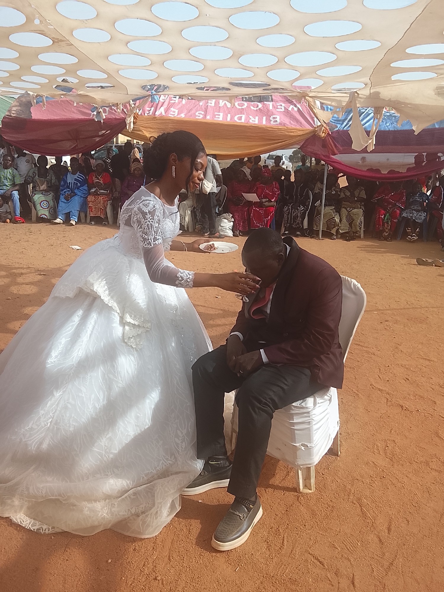 The image shows a wedding scene, likely in Dogon Dinia, Nigeria. A bride in a white gown offers a drink from a glass to a man sitting on a white plastic chair. The man is wearing a dark suit. Behind them, a crowd of people is seated, observing the event. A canopy is visible overhead, providing shade. The ground appears to be sandy. A sign with the words "BIRDIE'S WELCOME EVERY" is partially visible on the canopy.