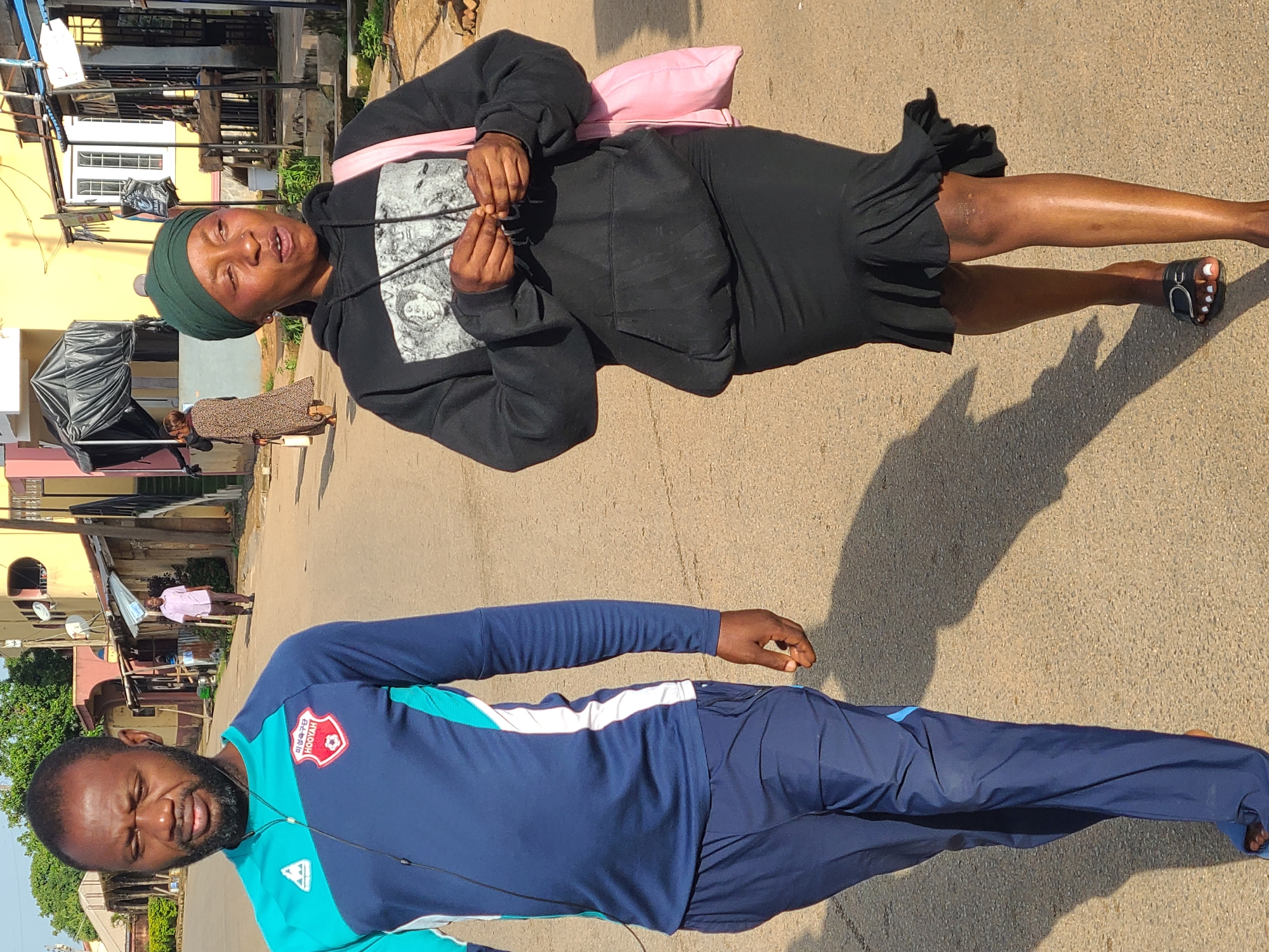 This media file captures an outdoor scene in Jos, Nigeria, likely during daylight hours given the strong shadows. The foreground features a man and a woman. The man is wearing a blue and teal long-sleeved athletic shirt with white accents and dark blue track pants. He has a beard and appears to be speaking or reacting to something, with his mouth open and eyes partially closed. He is also wearing earphones.
The woman is standing behind and to the left of the man, her head tilted back as if speaking or reacting. She is wearing a dark hoodie with a graphic print on the front and a ruffled skirt. A pink bag is slung over her shoulder. She has on a green headscarf and sandals.
In the background, there are buildings, some appearing residential, with visible architectural details like windows and balconies. A person in a pink shirt is visible walking further down the street. The ground is paved, suggesting a street or pathway. The overall impression is a candid street scene, possibly a casual encounter or conversation between the two individuals. The time of day appears to be morning or afternoon, indicated by the bright sunlight and distinct shadows. There is no discernible text visible in the image.