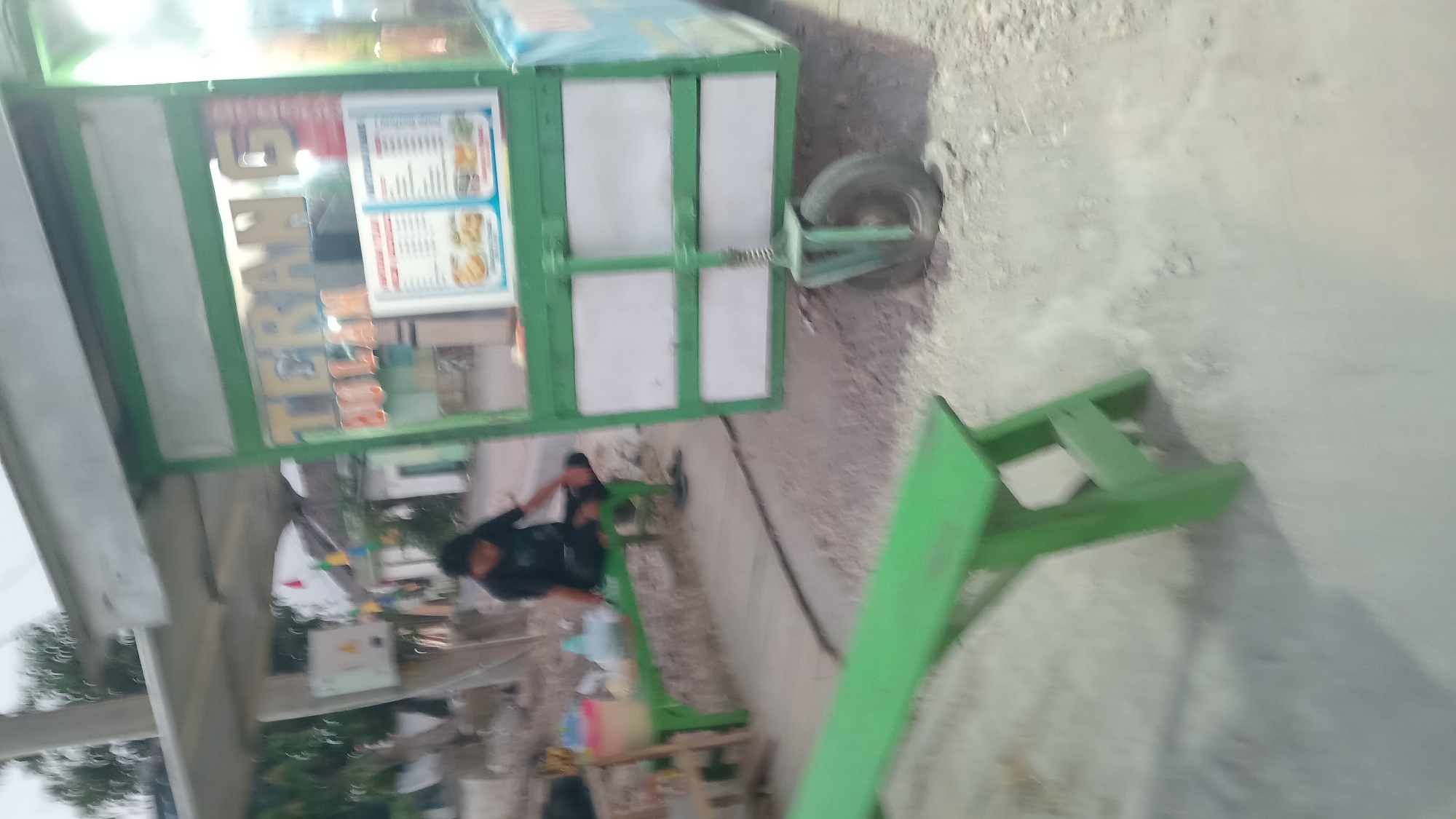 The image depicts an outdoor scene in Trembulrejo, Indonesia, featuring a food cart. A person is seated on a green bench to the left of the cart. The cart itself has a sign with text that appears to be "TERANG" and "BULAN". A menu is displayed on the front of the cart, showing images of food items and text. The cart has a green frame and white panels. A wheel is attached to the lower part of the cart. In the background, buildings and trees are visible. A second green bench is positioned in the foreground, to the left. The ground is covered with gravel and dust.