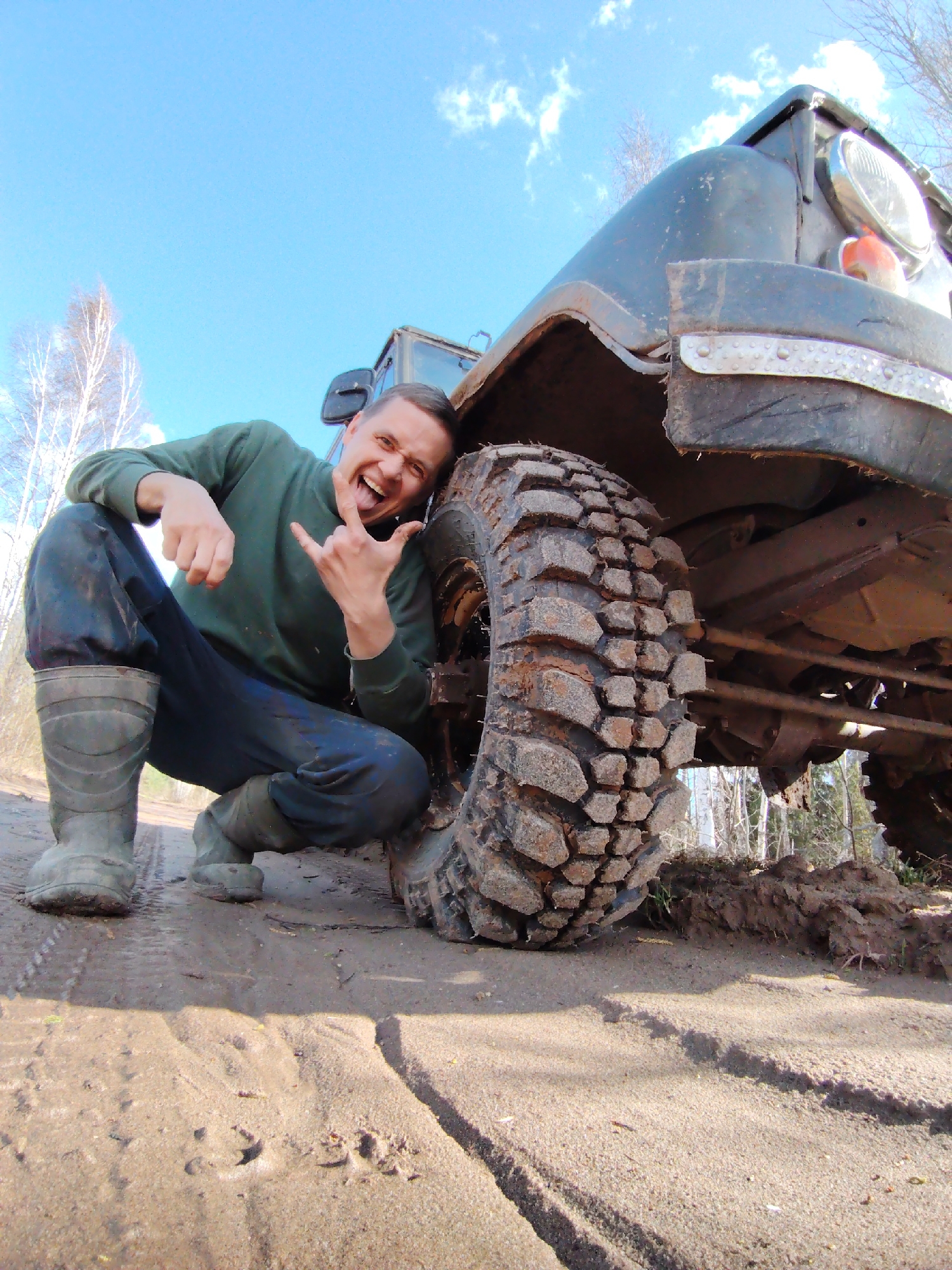 A man poses enthusiastically next to a rugged off-road vehicle on a sandy and muddy track in Markovskii, Russia. The man is crouching, leaning against the vehicle's front fender, and is captured from a low-angle perspective. He wears a dark green long-sleeved shirt, dark pants, and muddy rubber boots. With a wide, excited grin and his tongue playfully extended, he makes a "rock on" hand gesture with his left hand, conveying a sense of triumph or excitement.
The vehicle is an older, heavy-duty off-road utility vehicle, likely a UAZ, with a dark, mud-splattered exterior. Its large, deeply-treaded tire is prominent, caked with mud and dirt, suggesting it has been driven through challenging terrain. Visible elements of the vehicle include a round headlight, an amber turn signal, and a metal bumper. The immediate foreground shows a sandy track with tire impressions and areas of disturbed earth and mud.
The scene is set outdoors under a bright blue sky with a few wispy white clouds, indicating daytime and clear weather. In the background, sparse trees with bare branches are visible, characteristic of early spring or late autumn. The overall impression is that of an adventurous off-roading experience. No visible text is present in the image.