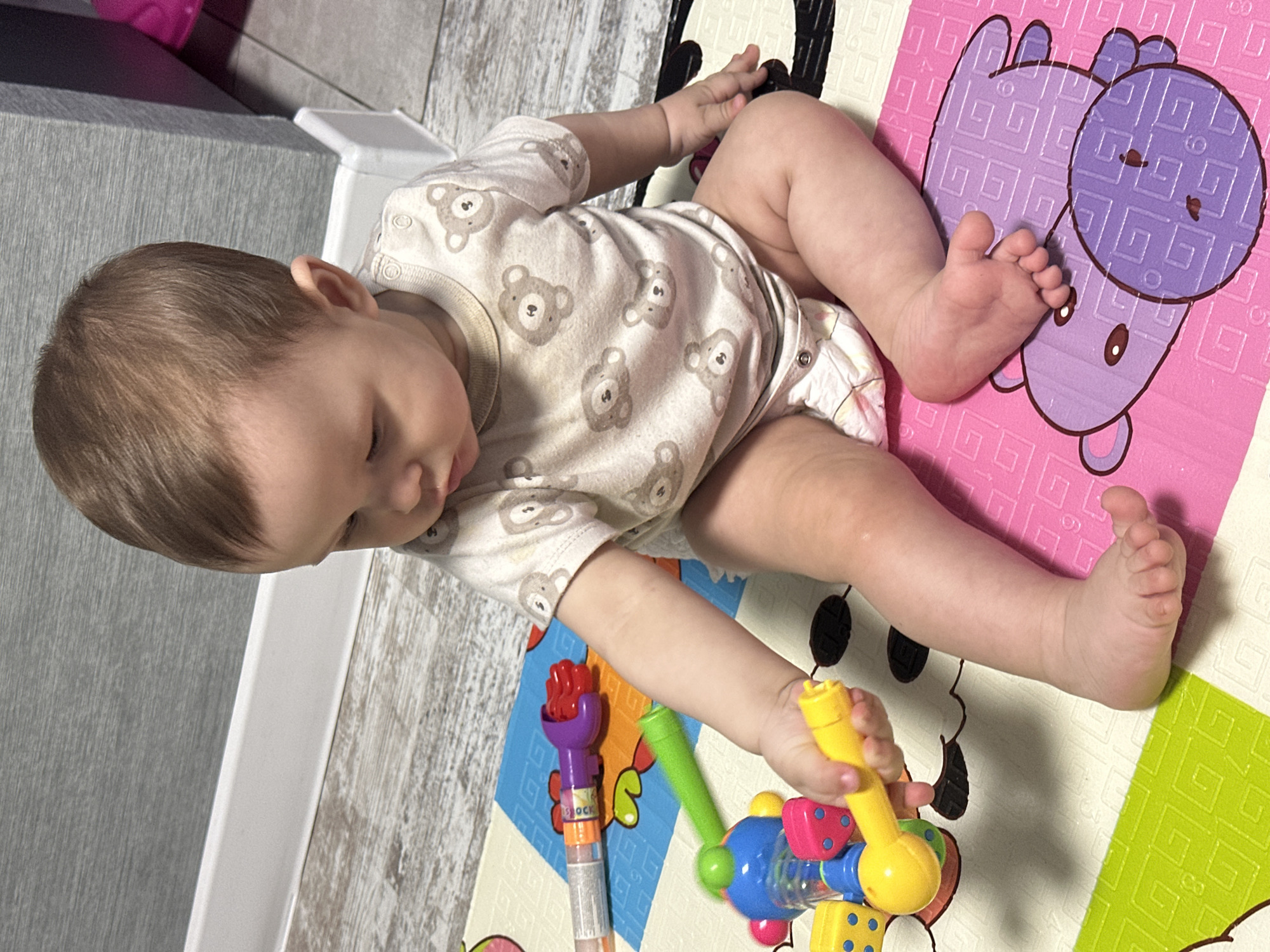 A fair-skinned infant, with light brown hair, is seated on a multi-colored foam playmat indoors in the city of Unknown, Unknown. The infant is wearing a light-colored, short-sleeved onesie patterned with numerous brown teddy bears, displaying a minor dark stain on the upper chest. A white disposable diaper is visible beneath the onesie. The infant holds a yellow plastic toy in their right hand, directing their gaze downwards towards it. Other plastic toys, including a purple grabber with a red claw, a green stick-like object, and a multi-colored shaker with blue, yellow, and pink components, are scattered on the playmat to the infant's left. The playmat consists of interlocking foam tiles featuring different colors and patterns, such as a purple section with a bear illustration, green with a grid pattern, and blue with geometric shapes. The floor surrounding the playmat is light-colored laminate or wood. White baseboards separate the floor from the light grey, textured wall in the background. A dark grey vertical structure and a partial view of a pink object are visible in the upper right background. The infant's bare feet are visible, with the right foot resting on the pink section of the playmat.