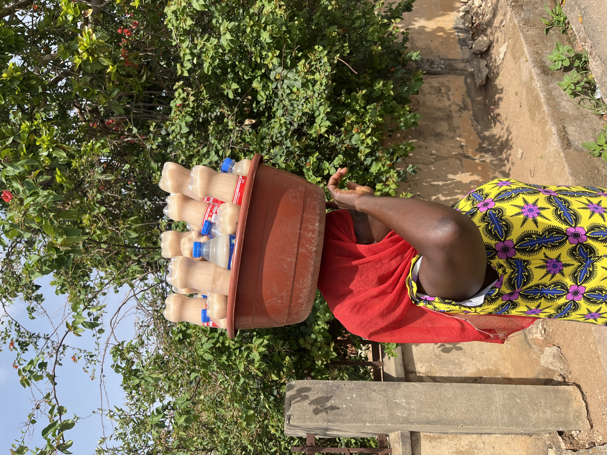 A woman in Jos, Nigeria carries a brown basin filled with approximately 15 plastic bottles of a light-colored liquid on her head. She is wearing a red head covering and a yellow and purple patterned garment. She is positioned next to a concrete ledge or low wall with greenery in the background. The setting appears to be outdoors in daylight.