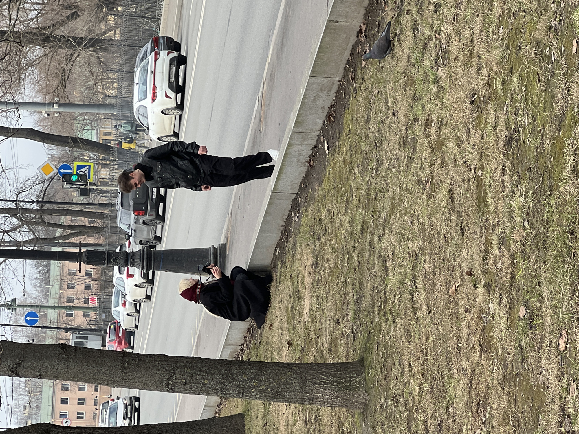 The image shows a street scene in St. Petersburg, Russia. A man in a black jacket and black pants with white sneakers is walking along a sidewalk. A woman wearing a black cloak and red head covering is sitting down taking photos. A pigeon is on the grass. Several cars are parked on the street. A traffic light is visible, along with street signs and a metal fence. A tree is in the foreground. Buildings are visible in the background.