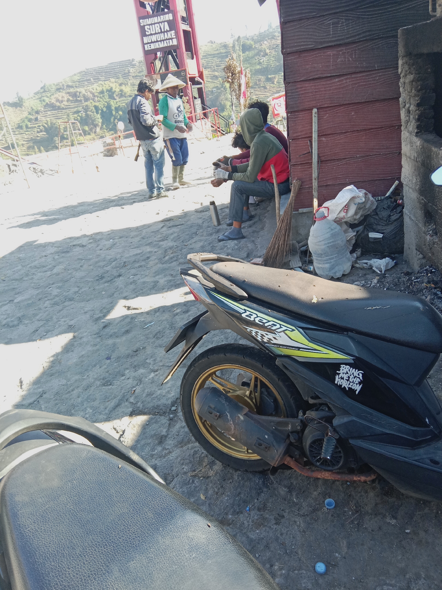 In the village of Tieng, Indonesia, an outdoor scene unfolds under a bright sun. In the foreground, a black scooter with gold accents is parked. Behind it, to the left, a man wearing a conical hat and green shirt stands talking to another man in a grey jacket and jeans. In the background, a red tower-like structure with signage in Indonesian is visible. A group of people are seated to the right, near a wooden building. The ground is covered in gravel and dirt, with some discarded items and a broom leaning against the building. Terraced hillsides with green vegetation rise in the distance. The overall impression is one of a casual, perhaps tourist-oriented, outdoor gathering in a rural Indonesian setting.