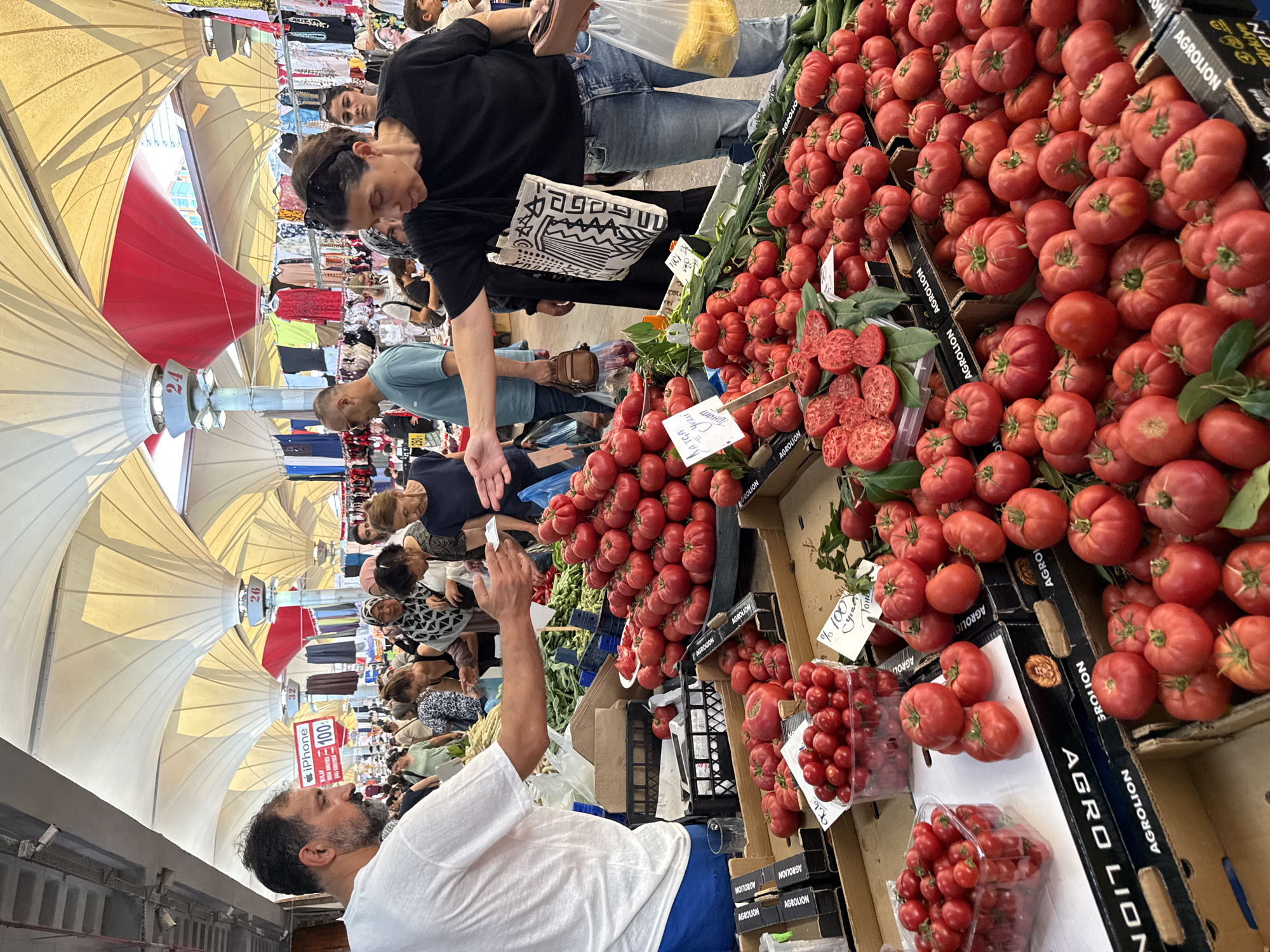The image captures a bustling scene at a covered market in Kadıköy, Turkey, likely a farmers market. In the foreground, a vendor wearing a white t-shirt and blue shorts is interacting with customers, holding out what appears to be money or a receipt. Directly in front of him, several crates and containers are overflowing with vibrant red tomatoes of various sizes, including a clear plastic container filled with cherry tomatoes. To the right, a woman in a black t-shirt and jeans is reaching out, possibly to select tomatoes or complete a transaction. She has a tote bag slung over her shoulder with a distinctive black and white pattern. Behind the central figures, the market extends with more stalls displaying produce, clothing, and other goods. The market is covered by a unique canopy of large, white, sail-like structures supported by poles, with red fabric accents visible in the background. Shoppers mill about, browsing the offerings. The overall atmosphere is one of lively commerce and community gathering within the market setting.
