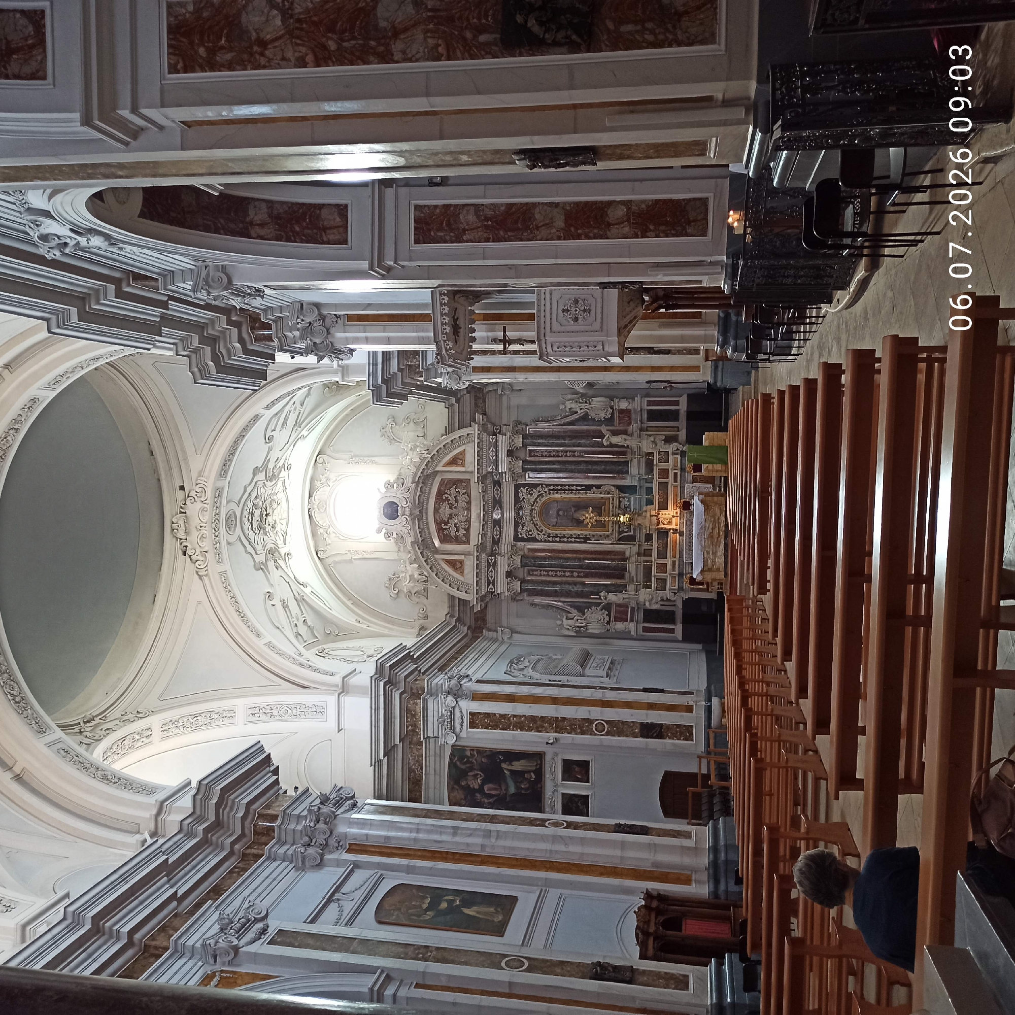 This image shows the interior of the Basilica di Santa Maria Assunta in Ruvo di Puglia, Italy. The camera is positioned facing the altar, with rows of wooden pews filling the foreground and mid-ground. A single person, seen from behind, is sitting in one of the pews on the left side of the frame. The architecture of the basilica is grand, with high vaulted ceilings, ornate decorations, marble columns, and large paintings adorning the walls. The altar area is elaborate, featuring a central painting, sculptures, and candles. The overall lighting suggests it is daytime, with light streaming in from a large window above the altar. The date and time stamp "06.07.2026 09:03" is visible in the bottom right corner.