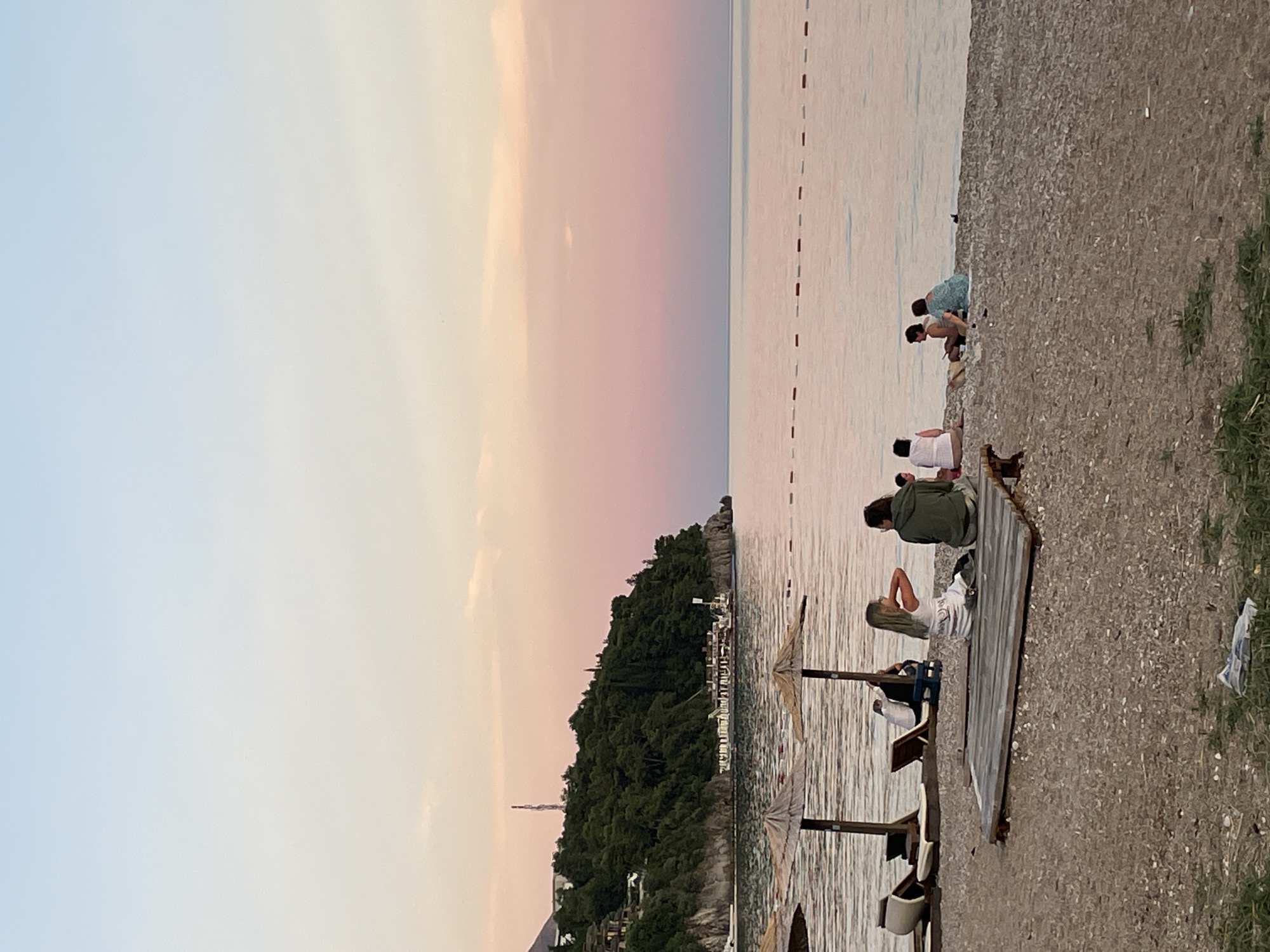 The image depicts a serene beach scene at dusk in Budva, Montenegro. The sky is a gradient of soft blues and pinks, indicating the sun is setting. The sea is calm, with gentle ripples reflecting the pastel colors of the sky.
Several people are scattered along the pebble beach and near the water's edge. On the beach, a group of individuals are seated on the shore, some facing the water. Further down, a person is leaning back on a wooden structure, possibly a pier or deck, looking out towards the sea. Another individual is sitting on the same structure, also facing away from the camera. The people appear relaxed, enjoying the peaceful atmosphere.
Objects visible include beach umbrellas, one of which is open, providing shade. There are also some lounge chairs. The beach itself is covered in small pebbles and some patches of dry grass are visible on the right side of the frame. In the background, a lush green hillside covered in trees slopes down towards the water. Some buildings and structures are nestled into the hillside, suggesting a coastal town. A tall, slender tower is also visible on the hill. The overall mood is tranquil and inviting. The time of day is clearly evening, as evidenced by the sky colors and the soft lighting. The location cues, such as the coastal landscape and architecture, are consistent with Budva, Montenegro.