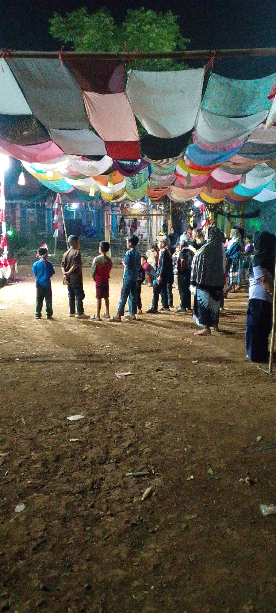 In a scene possibly located in Cilaku, Indonesia, an outdoor gathering is taking place under a canopy of colorful fabrics strung across a support structure. Several children, dressed in casual attire, stand facing away from the camera in a dirt area. To the right, a group of adults are also present, some with their backs to the viewer. The background features dimly lit structures and vegetation. The ground is uneven and covered with dirt, scattered with a few pieces of litter. The overall lighting suggests it is evening or nighttime.