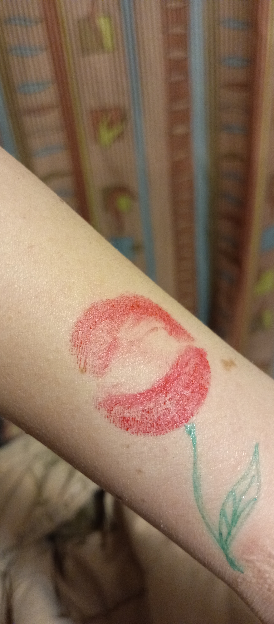 A close-up shot depicts a red lipstick imprint on a human arm, creatively transformed into a flower with a green stem and leaf drawn beneath it. The arm is lightly complected, and the background features blurred vertical stripes in shades of beige, blue, red, and yellow.