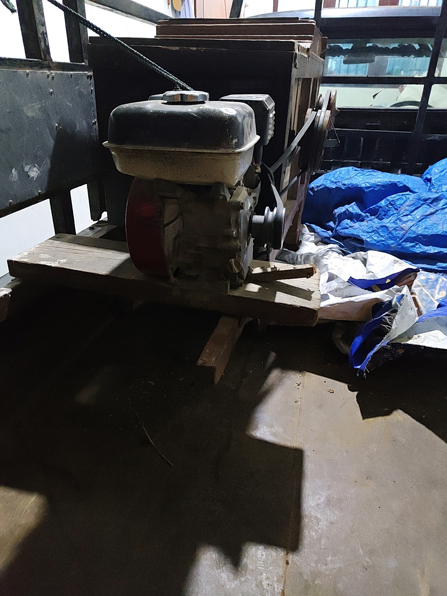 The image shows a close-up view of a disassembled or partially constructed wooden structure with a single-cylinder engine. The engine is black and silver, with a red fan shroud. It is mounted on a wooden platform supported by smaller wooden beams. To the right of the engine, a blue tarp is visible, partially covering some white and blue plastic sheeting. The background includes parts of a metal frame and windows, suggesting an outdoor or partially enclosed space. The lighting is dim, casting strong shadows, and the ground appears to be concrete. There are no people visible in the image. The overall impression is one of a work-in-progress or a storage area for equipment. This scene is located in Hutatoruan 11, Indonesia.