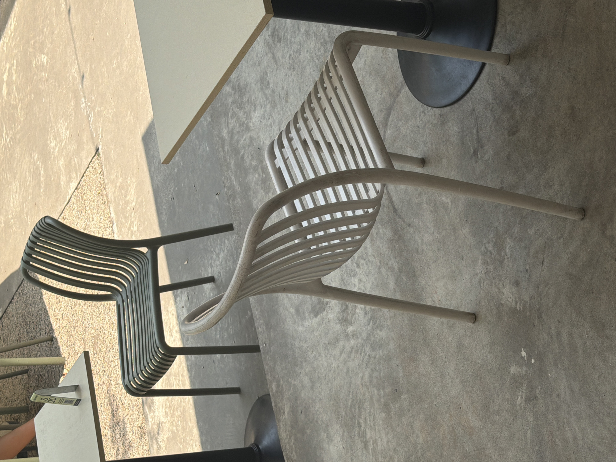 The image shows an outdoor seating area with two chairs and two tables on a concrete surface. One chair is light gray, the other is dark gray. One table has a light-colored square top, the other has a light-colored rectangular top. The location appears to be in daylight with a sidewalk area. An arm is visible near the lower-left corner of the image. The picture is probably taken in the city of Đà Nẵng, Vietnam.