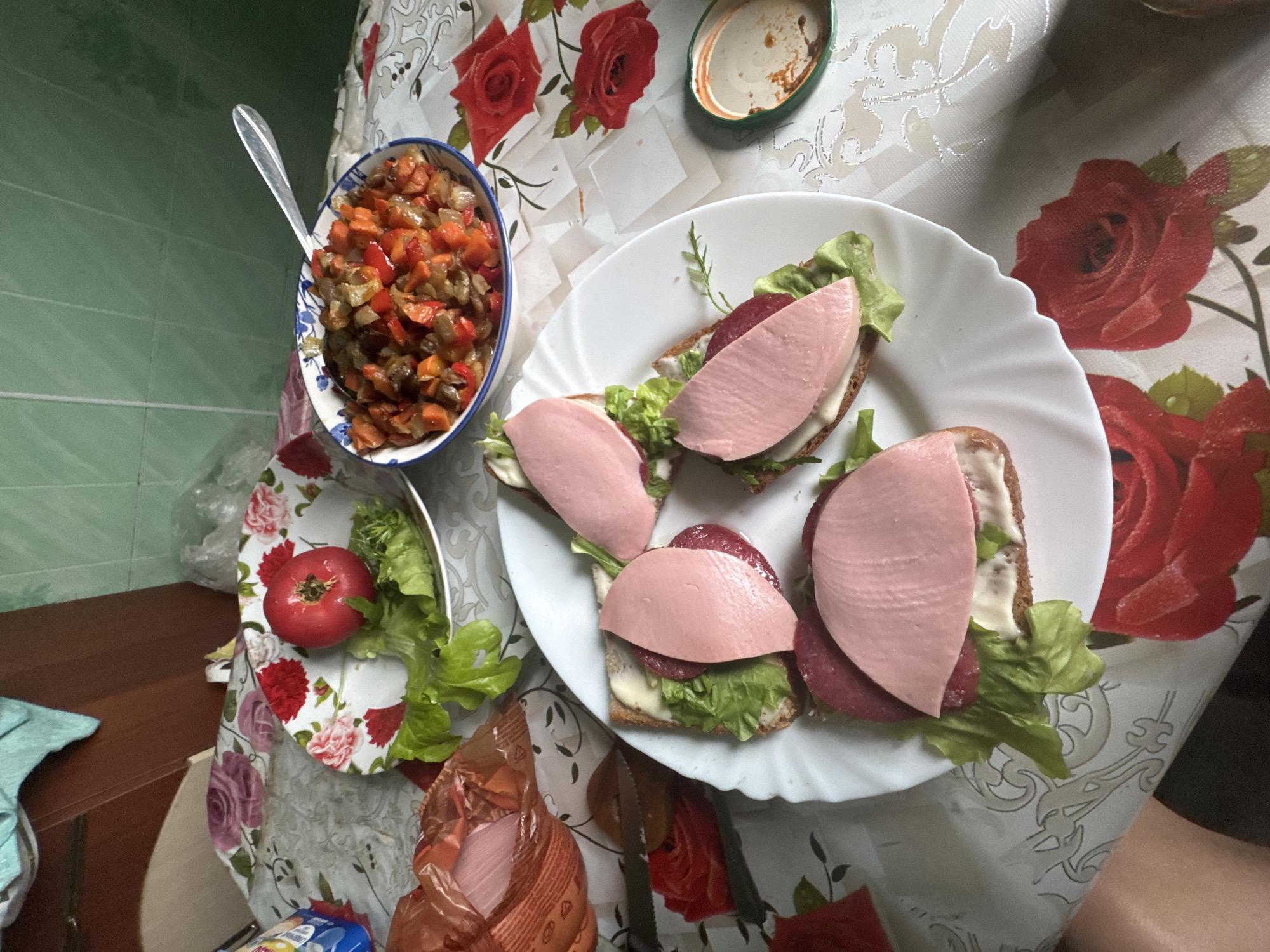 A top-down shot shows a meal laid out on a table covered with a floral tablecloth in Krasnoye, Russia. The table is set with a white plate of four open-faced sandwiches, each topped with lettuce, a slice of red sausage, and a thick slice of pink bologna. To the right of the sandwiches is a small green jar with a metal lid, and further to the right, a bowl of stewed vegetables including peppers, carrots, and onions. Behind the sandwiches, a white plate holds a single red tomato and a small pile of lettuce leaves. A bag of sliced bologna is partially visible on the left side of the table. The background shows a green tiled wall and wooden cabinets.