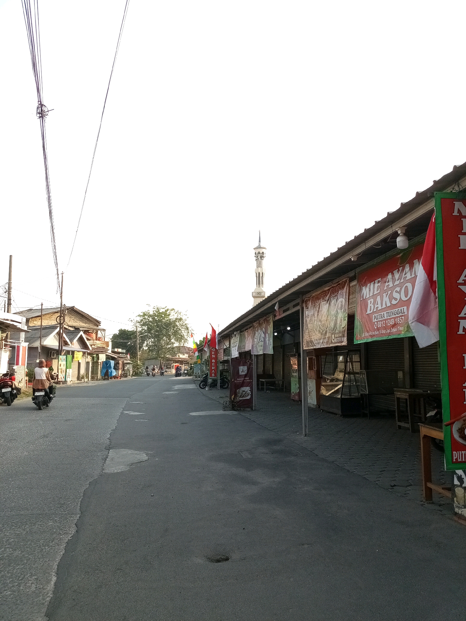 The image depicts a street scene in Bekasi, Indonesia, likely in a commercial area. A paved road runs through the center of the frame, with buildings lining the right side. These buildings appear to be shops or food stalls, indicated by the awnings and signage. One prominent banner displays "MIE AYAM BAKSO" in large text, along with contact information. A red and white Indonesian flag is also visible on the right side of the frame. The buildings on the left side of the road are simpler, residential or small commercial structures.
Several people are present. Two individuals are riding motorcycles on the left side of the road, moving away from the viewer. In the background, more motorcycles and individuals can be seen on the street, suggesting a moderate amount of activity. The sky is overcast and bright, dominating the upper portion of the image. A minaret of a mosque is visible in the distance, to the left of the shops. Overhead power lines cross the sky in the upper left quadrant. The overall impression is a typical street scene in a developing urban area, with a mix of commercial and residential elements.