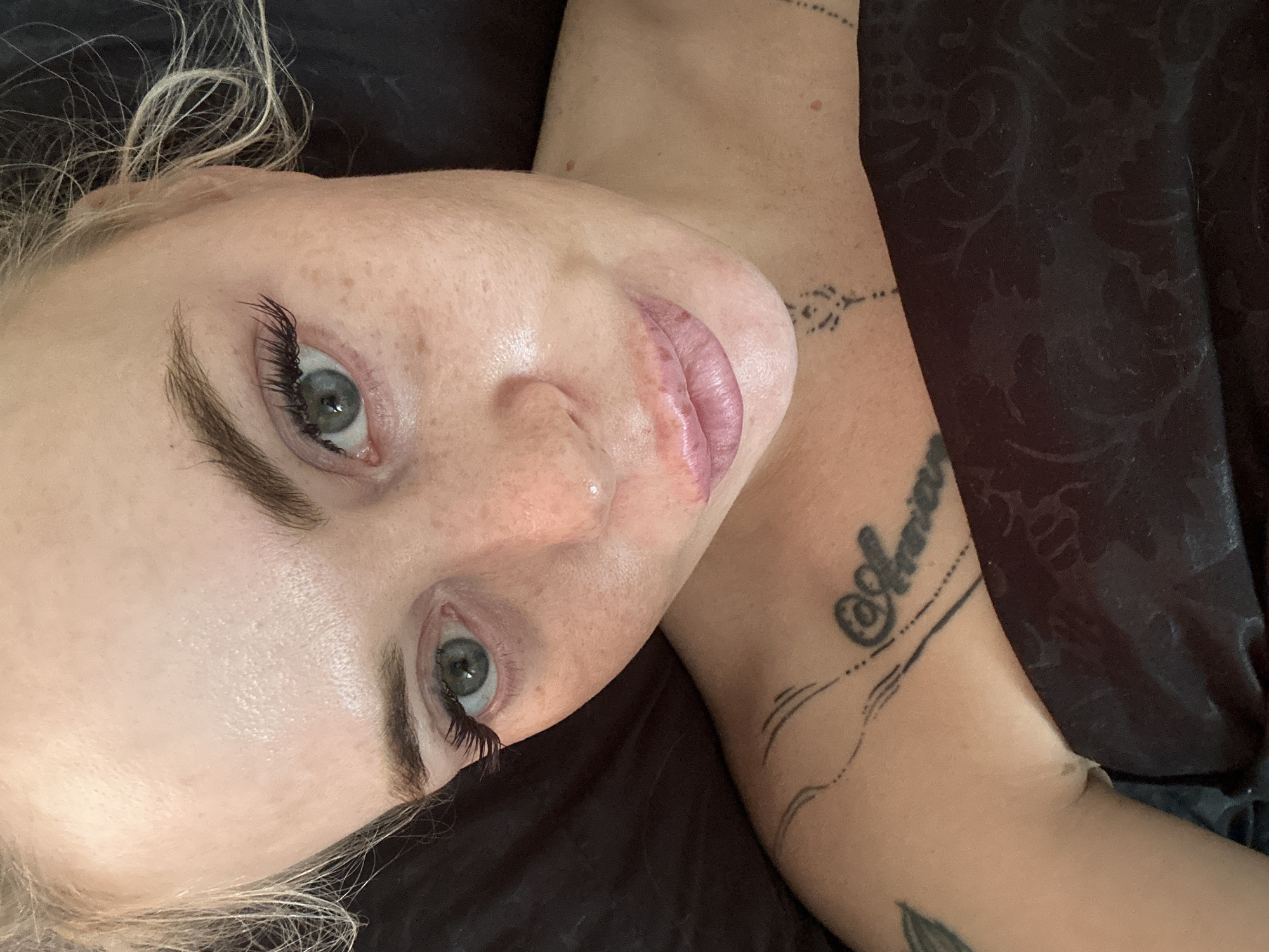 A close-up, overhead shot shows a fair-skinned woman lying down with her head tilted slightly. Her eyes are open and looking up. She has blue-gray eyes, dark eyebrows, and is wearing dramatic false eyelashes. Her face has visible freckles and some areas of redness around her nose and lips. Her lips are plump and appear to have been recently treated or are wearing lip gloss. She has blonde hair that is slightly messy. A dark, patterned fabric is visible covering her chest and shoulder. There are tattoos visible on her left shoulder and chest, including what appears to be the name "Annica" on her collarbone. The background is dark and out of focus. The location is Unknown, Unknown.