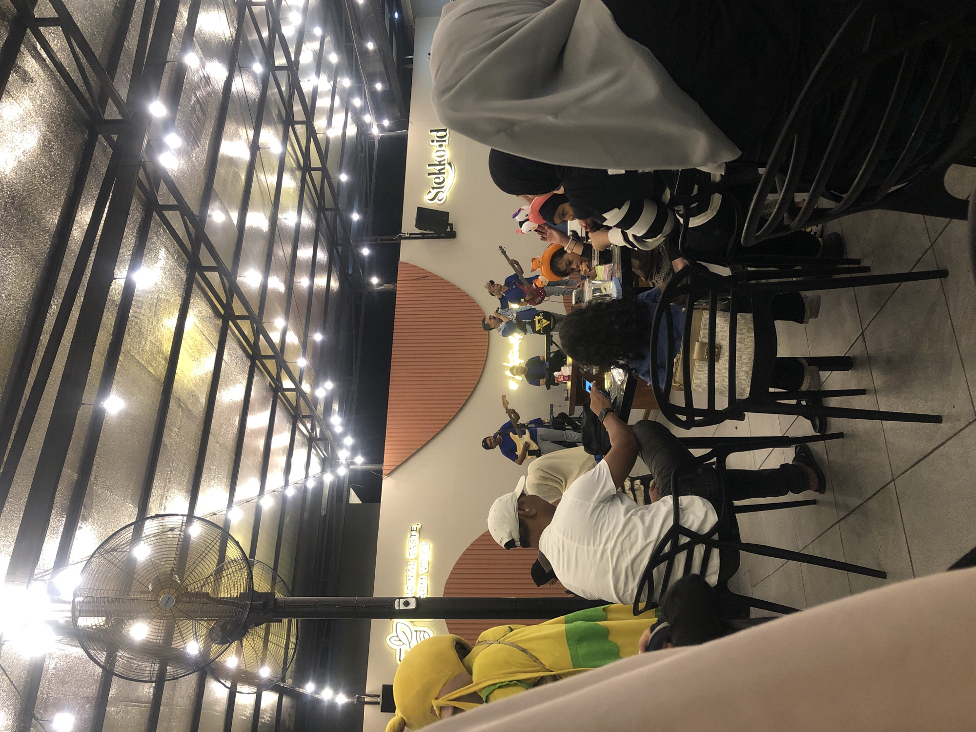 The image shows an indoor scene in what appears to be a restaurant or cafe. The location, based on the signage "Stekko-id" visible on the wall, is likely in Lhokseumawe, Indonesia. Several individuals are seated at tables. A live band or musical performance is taking place on a raised platform or stage, with members playing guitars and singing. The lighting consists of strings of decorative bulbs illuminating the ceiling, as well as neon signage. There is a large standing fan. Some patrons are looking at their phones, while others appear to be watching the performance. The overall environment seems to be casual and lively.