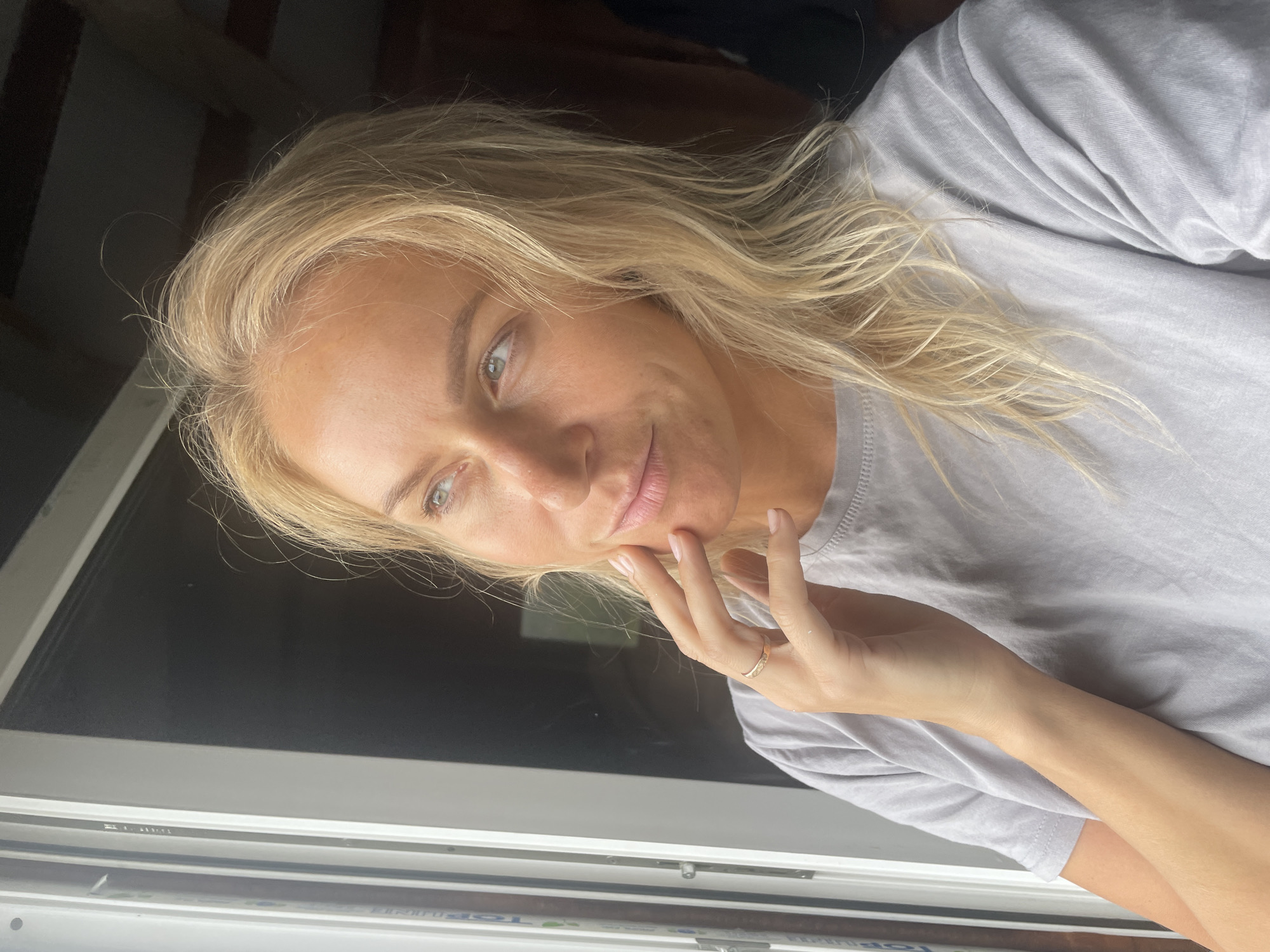 A woman with blonde hair and fair skin is posing near a window. She is wearing a gray t-shirt and a ring on her finger. Her hand is touching her face. The location is not discernible as Baranovka, Russia.