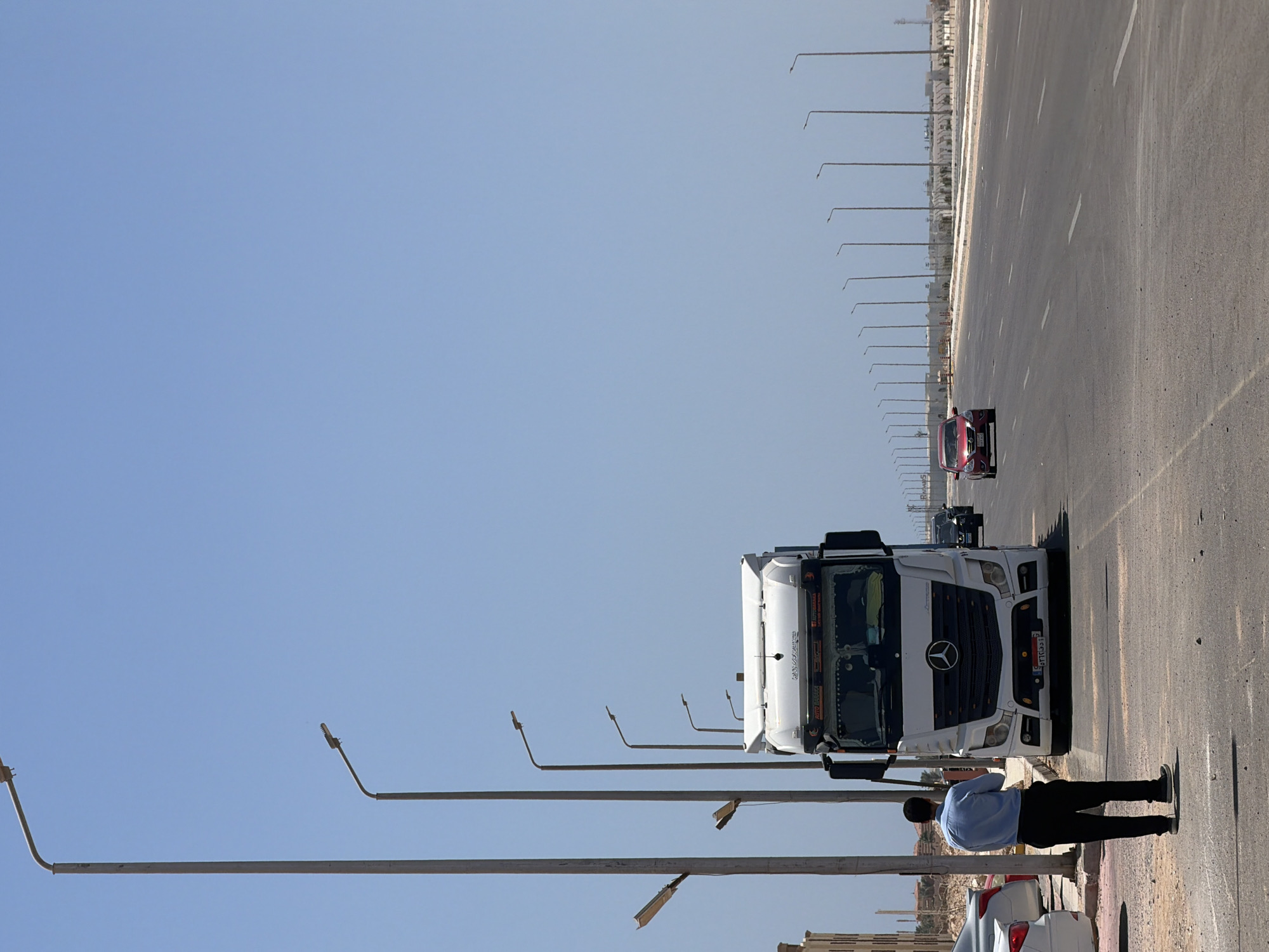 A man in a blue shirt and black pants stands on the side of a paved road, facing away from the camera and towards a white semi-truck. The truck, a Mercedes-Benz Actros, is parked on the right side of the road. Several other vehicles, including a red sedan and dark-colored SUVs, are visible on the road. The environment appears to be an urban or suburban area, with buildings and construction in the background. The sky is clear and blue, and the lighting suggests it is daytime. The scene is set in the city of Unknown, Egypt.