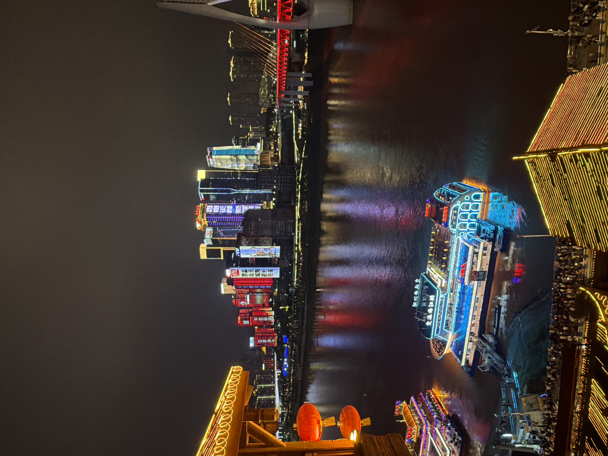 This is a nighttime photograph taken in Yuzhong District, China, likely from a vantage point overlooking a river and a cityscape. In the foreground, a traditional wooden structure with illuminated decorative lights is visible on the left, adorned with two red lanterns. Below this structure, two brightly lit tour boats are docked on the river. The boat on the left is smaller and appears to be a passenger ferry, while the boat on the right is a larger, multi-decked vessel with vibrant blue and green neon lighting. The river itself reflects the colorful lights of the city and the boats, creating a shimmering effect.
In the mid-ground, the wide river flows, separating the viewer from the opposite bank. The opposite bank is dominated by a dense cluster of modern skyscrapers, all illuminated with various colored lights, primarily red, white, and blue, creating a dynamic urban skyline. A prominent red suspension bridge with illuminated cables spans the river in the background, connecting the two sides of the city. The overall impression is one of a bustling and visually striking urban environment at night. The scene suggests a tourist or entertainment district, given the presence of the illuminated boats and the scenic waterfront.