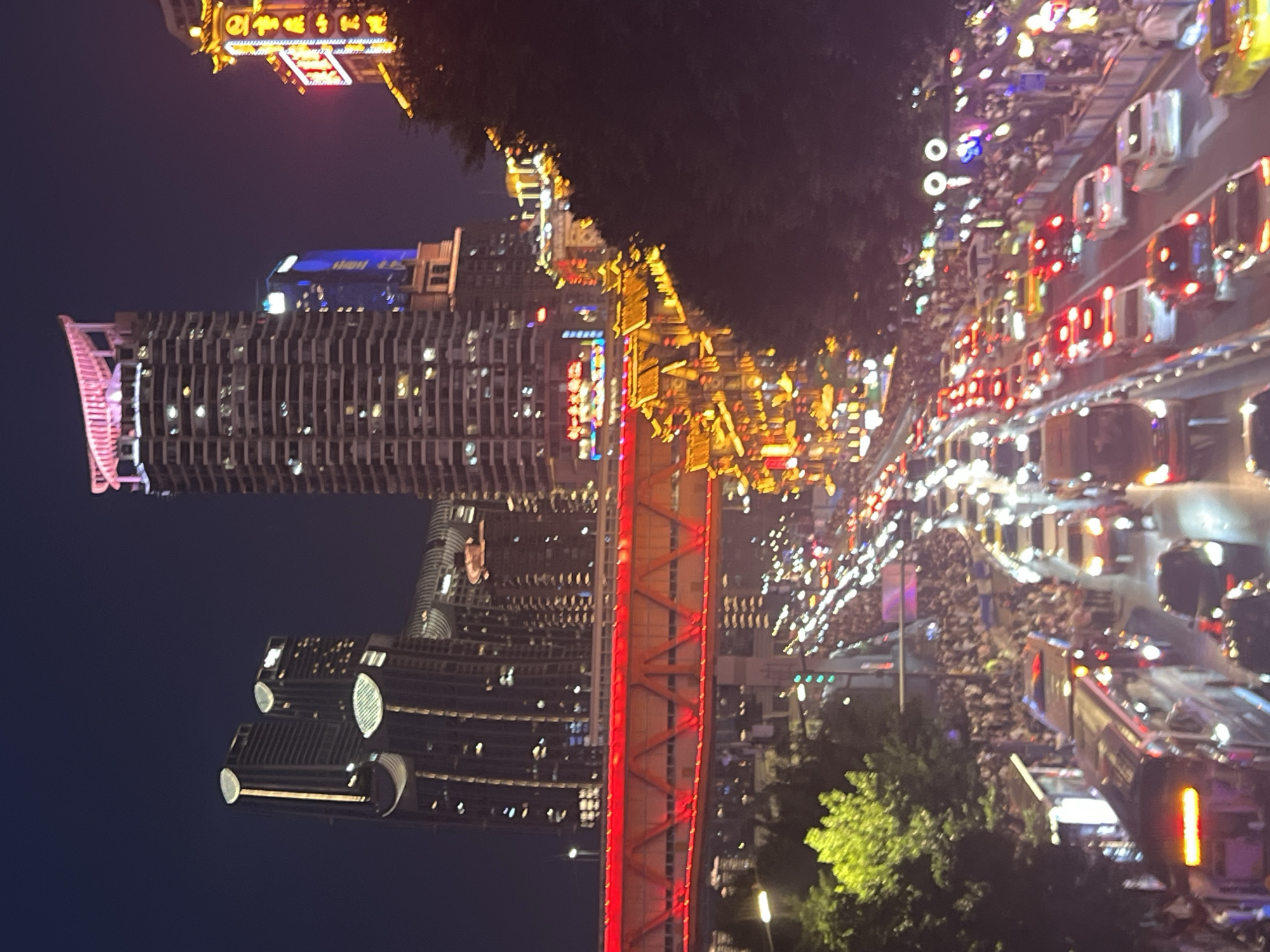 The image shows a bustling city scene at night, likely in the Yuzhong District of China, characterized by vibrant lights and a dense population. A multi-lane road is filled with a significant volume of traffic, with brake lights of numerous vehicles creating streaks of red illumination. Pedestrians are gathered in large numbers on sidewalks and possibly on the road itself, suggesting a crowded event or popular gathering spot.
Dominating the skyline are several tall buildings, including skyscrapers adorned with illuminated windows and decorative lighting, some featuring a pinkish glow at their apex. A distinctive red-lit bridge spans across the scene, adding a bright, artificial element to the nighttime environment. Below the bridge, a row of buildings with traditional East Asian architectural features, illuminated by warm golden lights, creates a contrast with the modern skyscrapers. Trees with lush green foliage are visible on the left side of the image, providing a natural element amidst the urban landscape. The overall atmosphere is one of energetic activity and urban illumination.