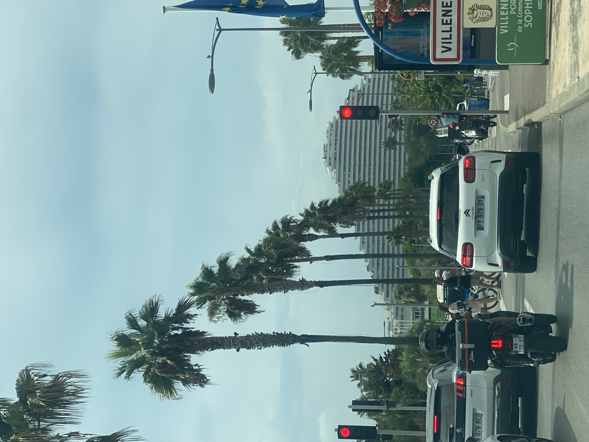 The image captures a street scene in Villeneuve-Loubet, France, likely during daylight hours under a cloudy sky. The foreground shows a traffic jam with several vehicles, including a motorcycle with a person wearing a helmet and a black jacket, a white Citroën SUV, and a white Peugeot SUV. Further ahead, a white tiered building, possibly a hotel or resort, with a distinctive architectural design dominates the background. Palm trees are prominently featured along the street, lining the road and creating a tropical atmosphere. Traffic lights are visible, with one red light illuminated. Signage for "Villeneuve-Loubet" is present, along with other directional or informational signs. People are visible on the sidewalk, including a woman and two men, one pushing a bicycle. A blue flag with yellow stars, likely the European Union flag, is seen on a flagpole to the right. The environment suggests a coastal or resort town.