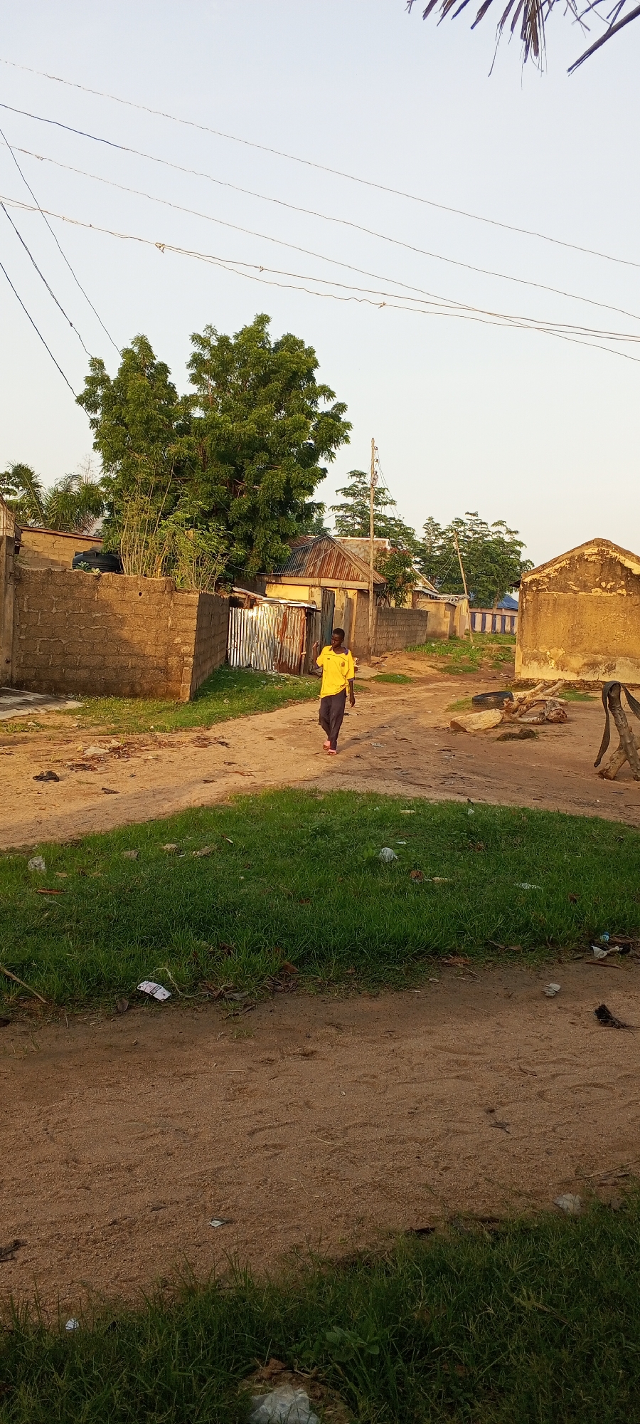 An outdoor, medium-shot photograph captures a young boy walking down a dirt road in Jalingo, Nigeria. He is wearing a bright yellow t-shirt and dark pants, with red shoes. The boy is walking away from the camera, toward a cluster of buildings in the background. The buildings appear to be made of mud brick and corrugated metal, and some have tin roofs. There is a large tree with green leaves to the left of the frame, and power lines stretch across the sky. The ground is a mix of dirt and patches of green grass. Scattered debris, including plastic bags and what appear to be old tires, are visible on the ground. The lighting suggests it is late afternoon.