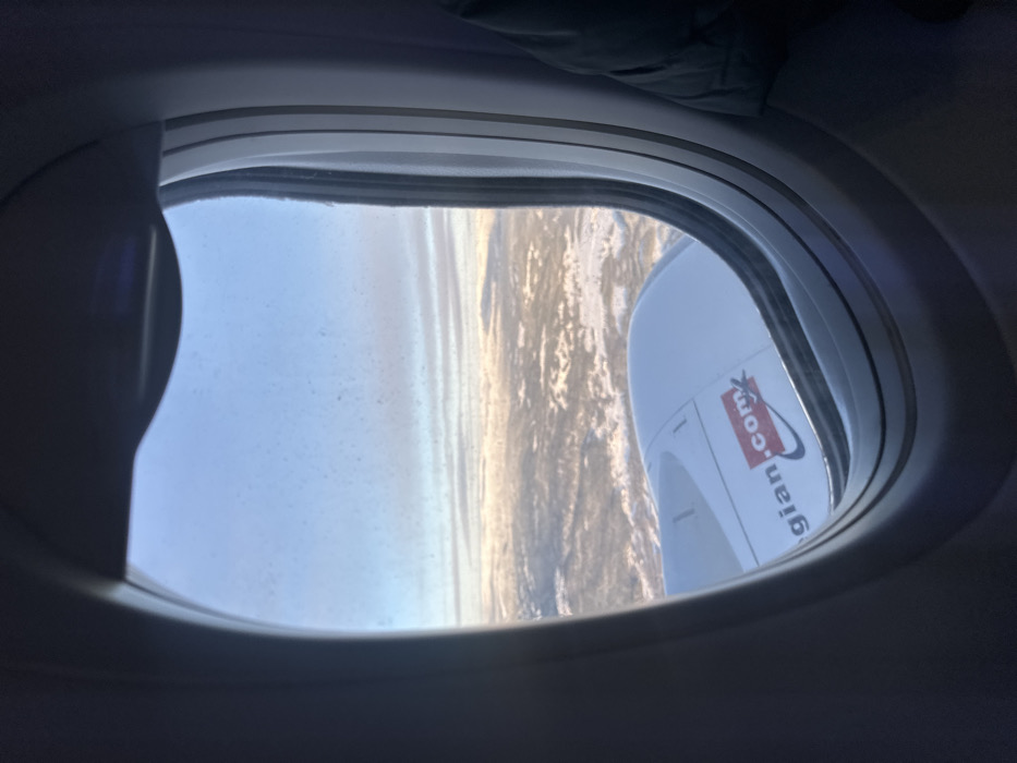 The image displays a view from an aircraft window, looking downward and forward over a vast, snow-covered landscape. The scene is observed from high altitude, with the ground below characterized by undulating terrain, partially covered in white snow, suggesting winter conditions. This landscape is situated in the city of Teigebyen, Norway.
The dark gray oval frame of the aircraft window occupies the immediate foreground. Through the lower left portion of the window, a segment of a white aircraft engine nacelle is clearly visible. A red and white logo, partially reading "Norwegian.com", is affixed to the exterior surface of this engine nacelle.
The sky above is a pale blue, transitioning to lighter tones near the horizon. Thin, stratified clouds are present in the distance. The outer surface of the aircraft window pane exhibits a scattering of small, dark specks, indicative of dust or particulate accumulation. No human figures or direct interactions are visible within the frame, though a dark garment or object is partially discernible on the far right, just inside the window frame, suggesting the presence of a passenger. The lighting indicates either early morning or late afternoon conditions, with soft, directional light.