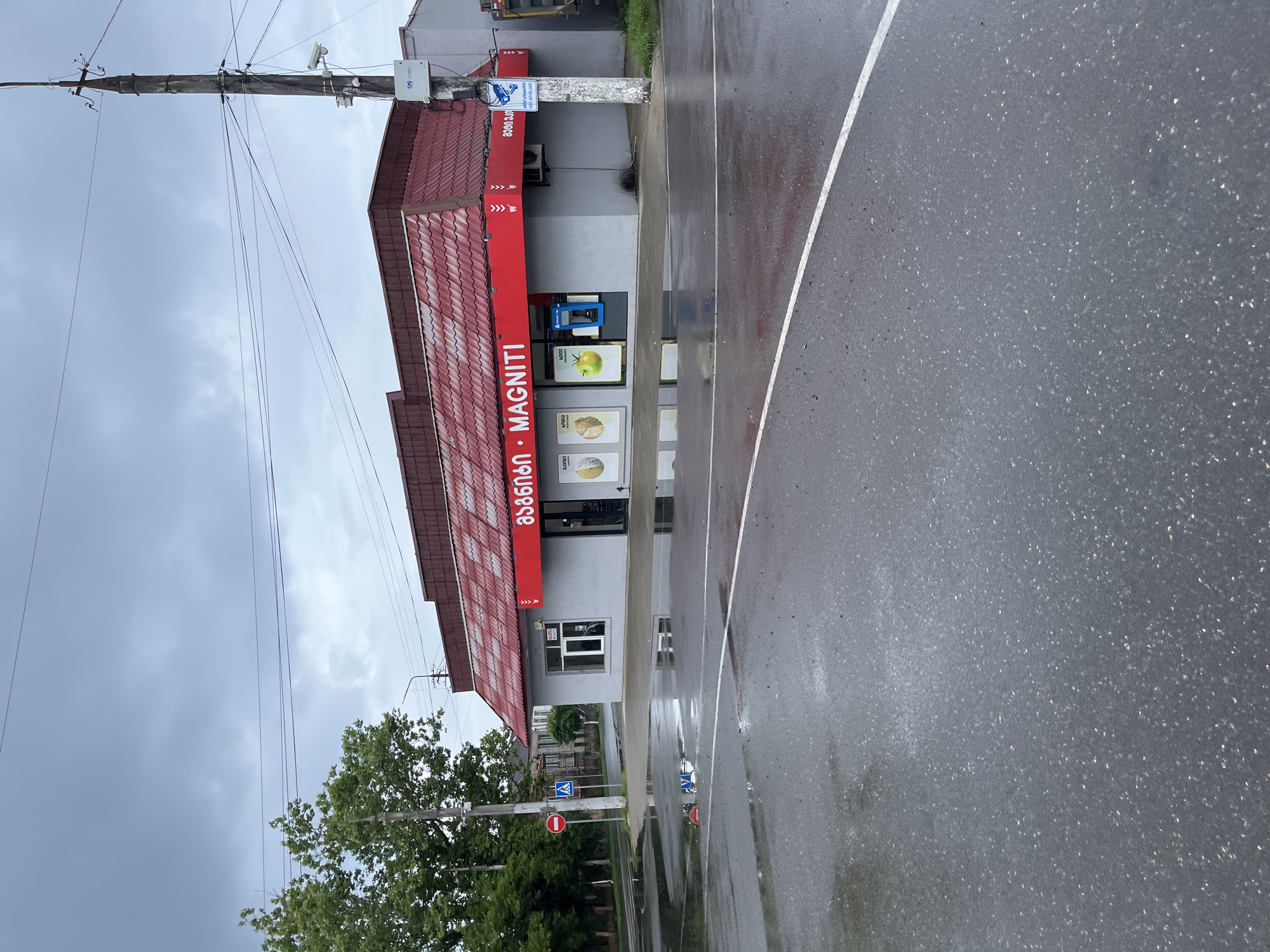 The image shows a low-angle, full shot of the Magniti store located in Poti, Georgia. The store, a single-story building with a red tiled roof, has a gray facade and a prominent red awning with white text that reads "MAGNITI" in Georgian and English. The store is situated on a street corner, with a wet asphalt road in the foreground, indicating recent rain. Puddles reflect the overcast sky and the building. To the left of the store, a traffic light and a pedestrian crossing sign are visible. A concrete utility pole stands to the right of the store, equipped with surveillance cameras and electrical wires. The sky is heavily overcast, with dark clouds suggesting a gloomy atmosphere. The overall scene depicts a commercial establishment in an urban setting, possibly after a period of rain.