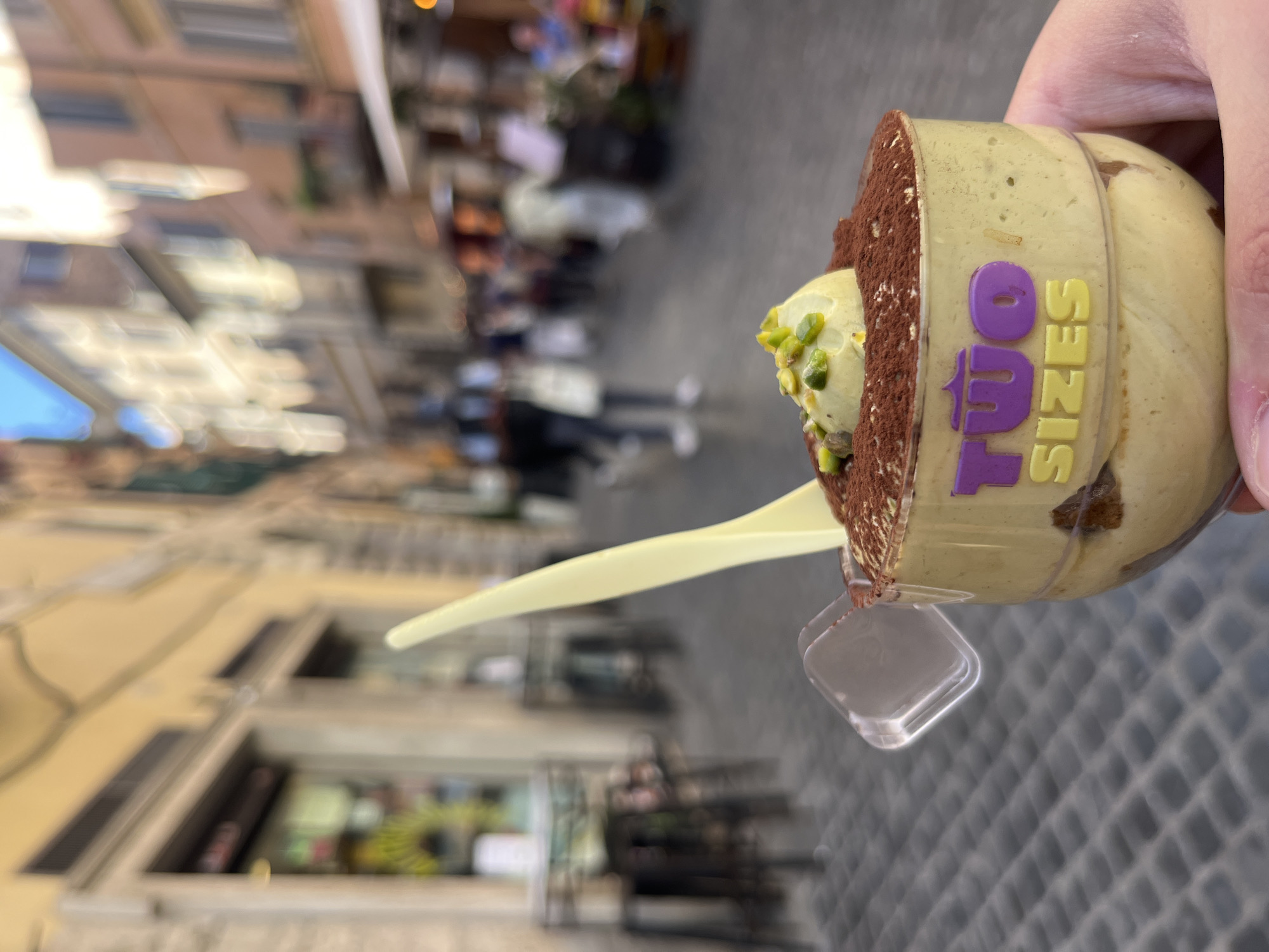 A close-up, slightly tilted photograph captures a hand holding a layered dessert against a blurred background of a bustling city street, identifiable as Rome, Italy. The dessert, presented in a clear plastic cup, features a generous topping of dark cocoa powder over a creamy, light green layer, likely pistachio-flavored, adorned with chopped pistachios. A pale yellow plastic spoon is inserted into the dessert, suggesting it's ready to be eaten.
A decorative band around the plastic cup prominently displays the text "TWO SIZES" in purple and yellow lettering. The hand holding the dessert is partially visible, showing a thumb and fingers.
The background is softly out of focus, depicting a vibrant daytime street scene. Buildings with visible windows line the street, and numerous blurred figures of people can be seen walking, hinting at a lively urban environment. The ground appears to be paved, consistent with city streets. The bright lighting suggests fair weather during the day. The image evokes a sense of enjoying a sweet treat amidst the atmosphere of a historic European city.