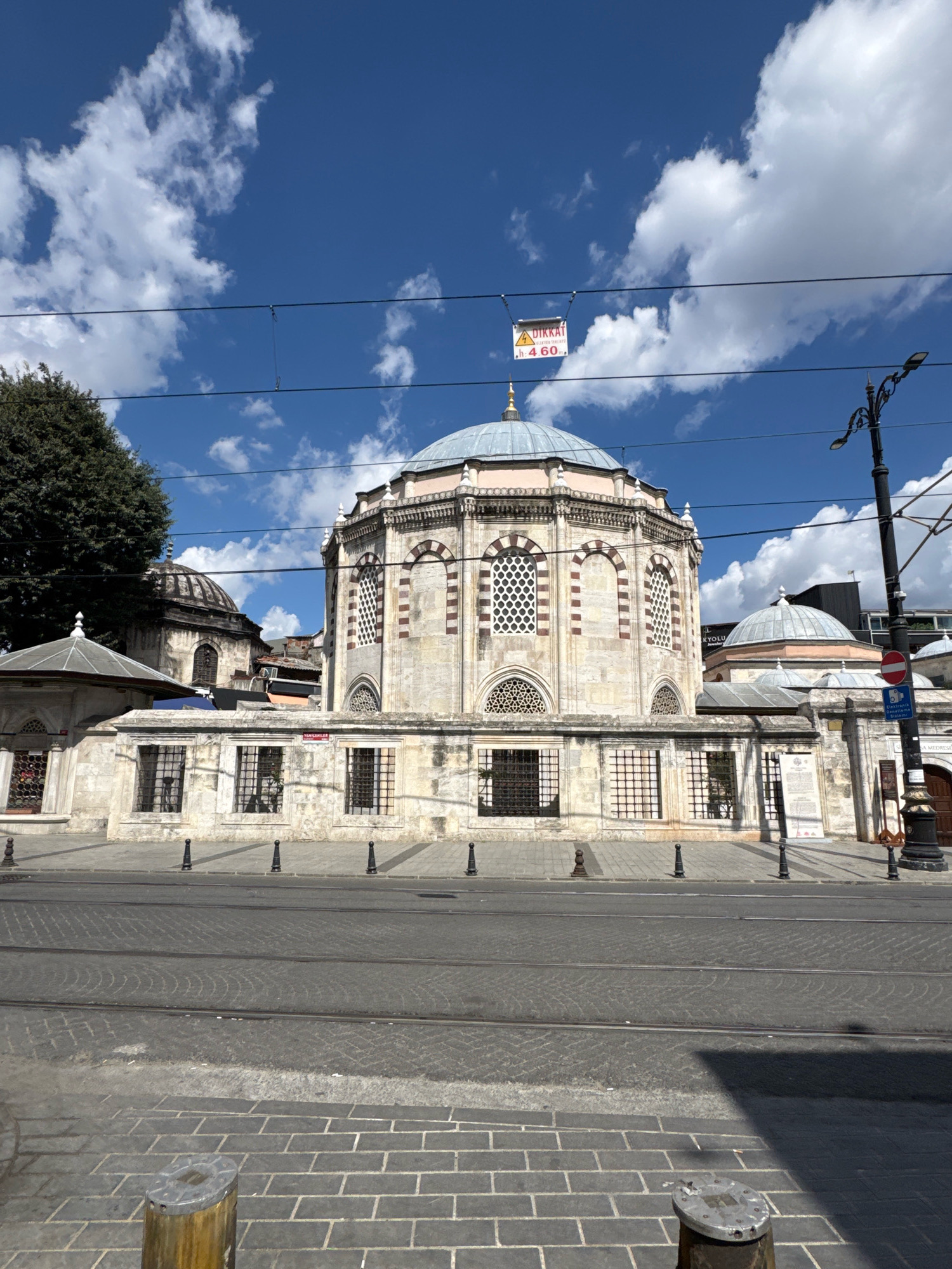 The image shows a view of the Eyüpsultan Mosque in Istanbul, Turkey, on a sunny day with scattered clouds. The main subject is the domed building of the mosque, constructed with stone and featuring arched windows and decorative bands of red and white stone. In front of the mosque is a low stone wall with barred windows, and beyond that, a street with tram tracks. To the left of the mosque, part of a large tree is visible. Overhead, power lines and a warning sign are suspended. A lamppost stands to the right of the frame. The foreground consists of a paved sidewalk and street.