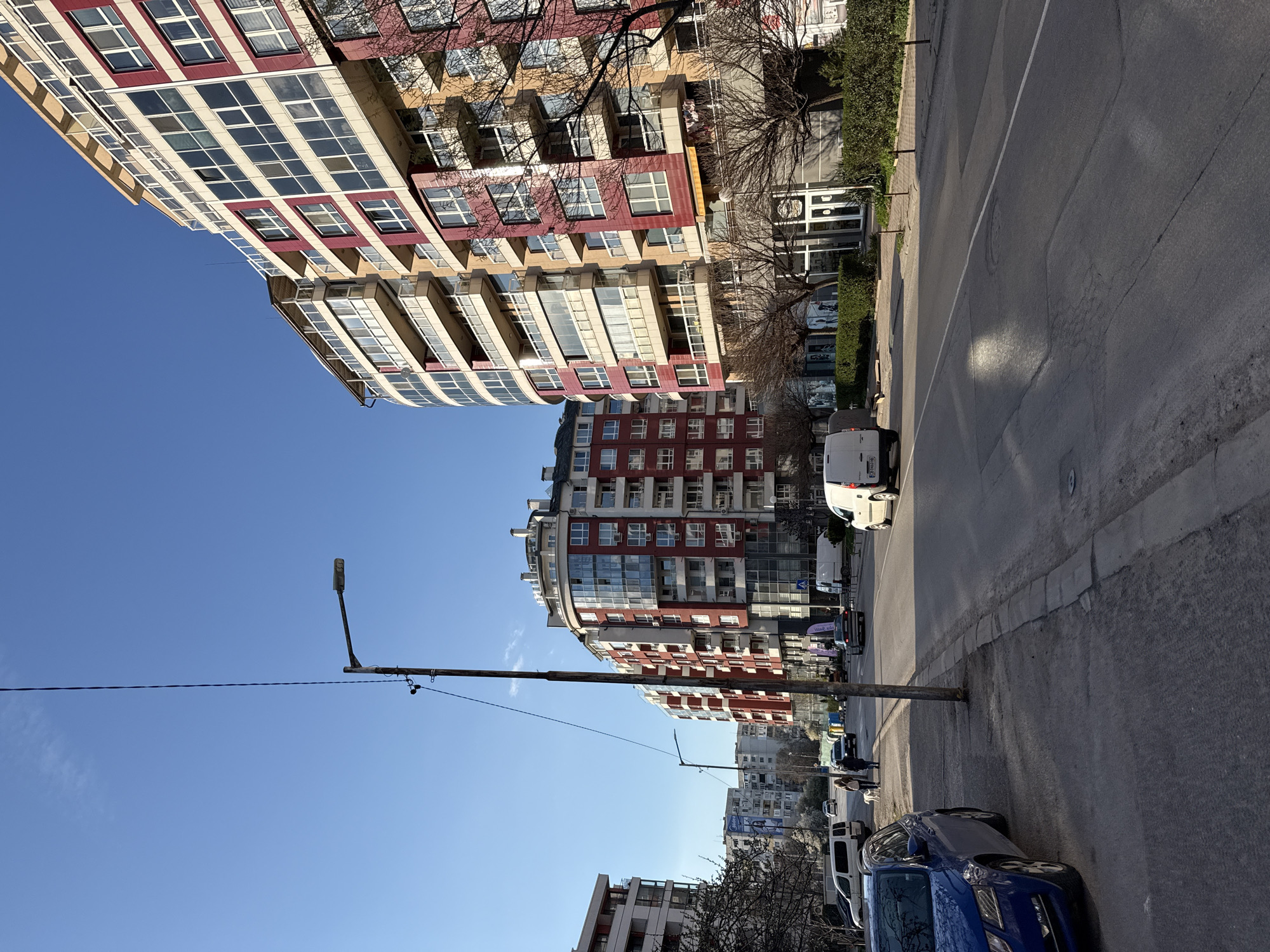 The image depicts a street scene in Sofia, Bulgaria. The photograph is taken on a street lined with modern apartment buildings, characterized by red and white facades and multiple balconies. Parked cars line the right side of the street. A white van is visible in the street. There are people walking, including a woman with a small white dog. Streetlights and overhead wires are present, against a clear blue sky.