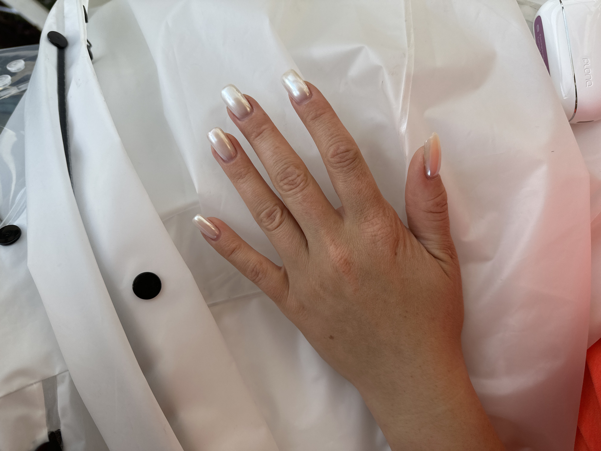 A hand with long, almond-shaped, pearlescent nails rests on a white, crinkled fabric, possibly a jacket or tarp. The hand is fair-skinned with visible veins and some freckles. The nails have a subtle pink and iridescent sheen, catching the light.
The setting appears to be indoors or under an overhang, as suggested by the bright, diffused lighting which creates soft shadows. The white fabric dominates the frame, with folds and creases creating texture. On the left side of the image, two black, circular snaps are visible on the white fabric, suggesting it is a garment. Part of another dark object, possibly another snap or a strap, is visible at the top left.
To the right of the hand, a white electronic device with a purple accent is visible. The brand name "plona" is legible on the device. The overall impression is a close-up shot focusing on the hand and nails against a neutral background. There are no explicit cues about the time of day, weather, or specific emotions being conveyed. The location context of Nizhny Novgorod, Russia, is not directly inferable from the visual elements alone.