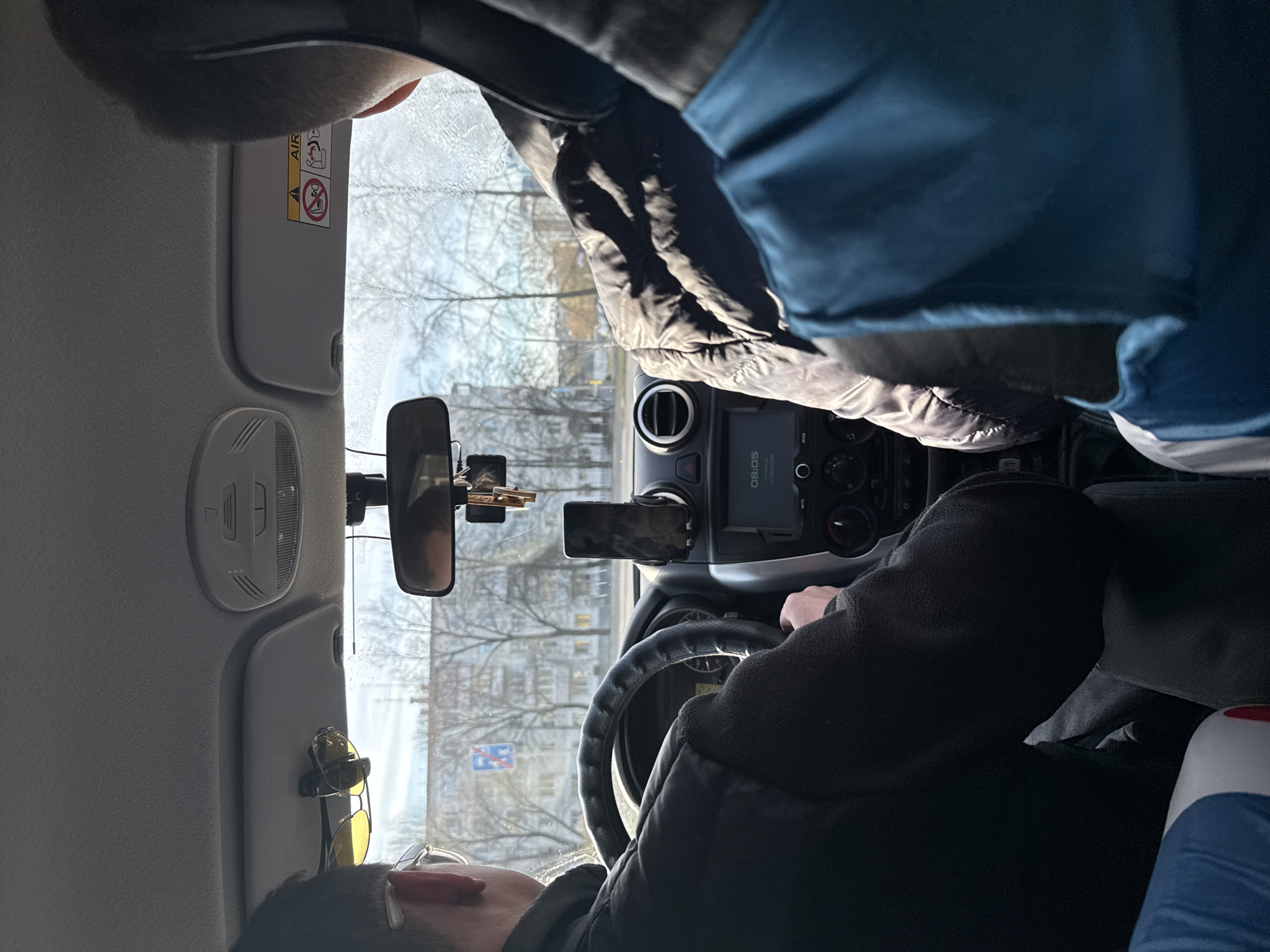 The image shows the interior of a car in Chaikovsky, Russia. The driver is partially visible, wearing glasses and a dark jacket. The car is on a road in the city; buildings and trees are visible through the windshield. A mobile phone is mounted on the dashboard. Keys are hanging from the rearview mirror. Sunglasses are clipped to the sun visor. A black jacket and a blue bag are on the passenger seat. The time displayed on the car's screen is 08:05.