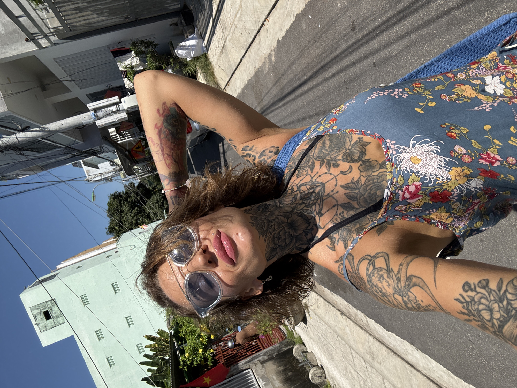 In Nha Trang Ward, Vietnam, a woman with extensive tattoos on her neck and arms is captured in a close-up selfie. She is wearing large, translucent sunglasses and a blue floral print dress, with a blue knitted cover-up visible. Her right arm is raised behind her head, and her left arm is extended, holding the camera. The background shows a street scene with buildings, power lines, trees, and a red flag. The lighting suggests it is daytime, with harsh shadows indicating bright sunlight.