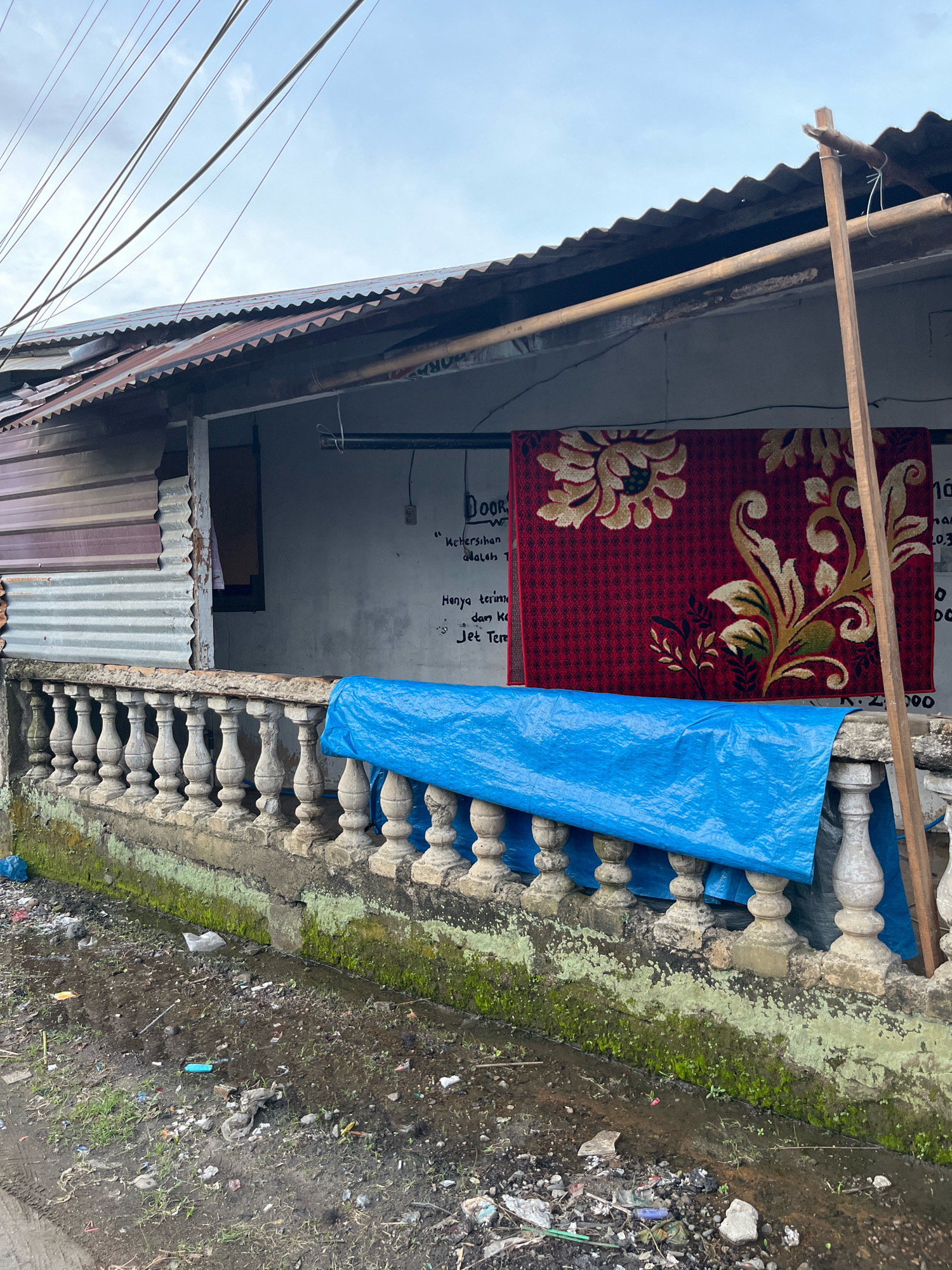 This image captures a scene on the street of Patumbak Dua, Indonesia. In the foreground, a muddy, debris-strewn ground shows signs of recent rain, with small puddles and scattered litter. A low, weathered concrete balustrade with ornate pillars runs horizontally across the middle of the frame. Above the balustrade, a vibrant red rug with intricate gold and green floral patterns is hung on a wall, partially obscuring a handwritten sign. A bright blue tarp is draped over the balustrade, casting a shadow. The background reveals the corrugated metal roof of a simple structure, supported by wooden poles. Overhead, a tangle of electrical wires crisscrosses the overcast sky, hinting at a utilitarian urban environment. The overall impression is one of humble living conditions within a densely populated area.