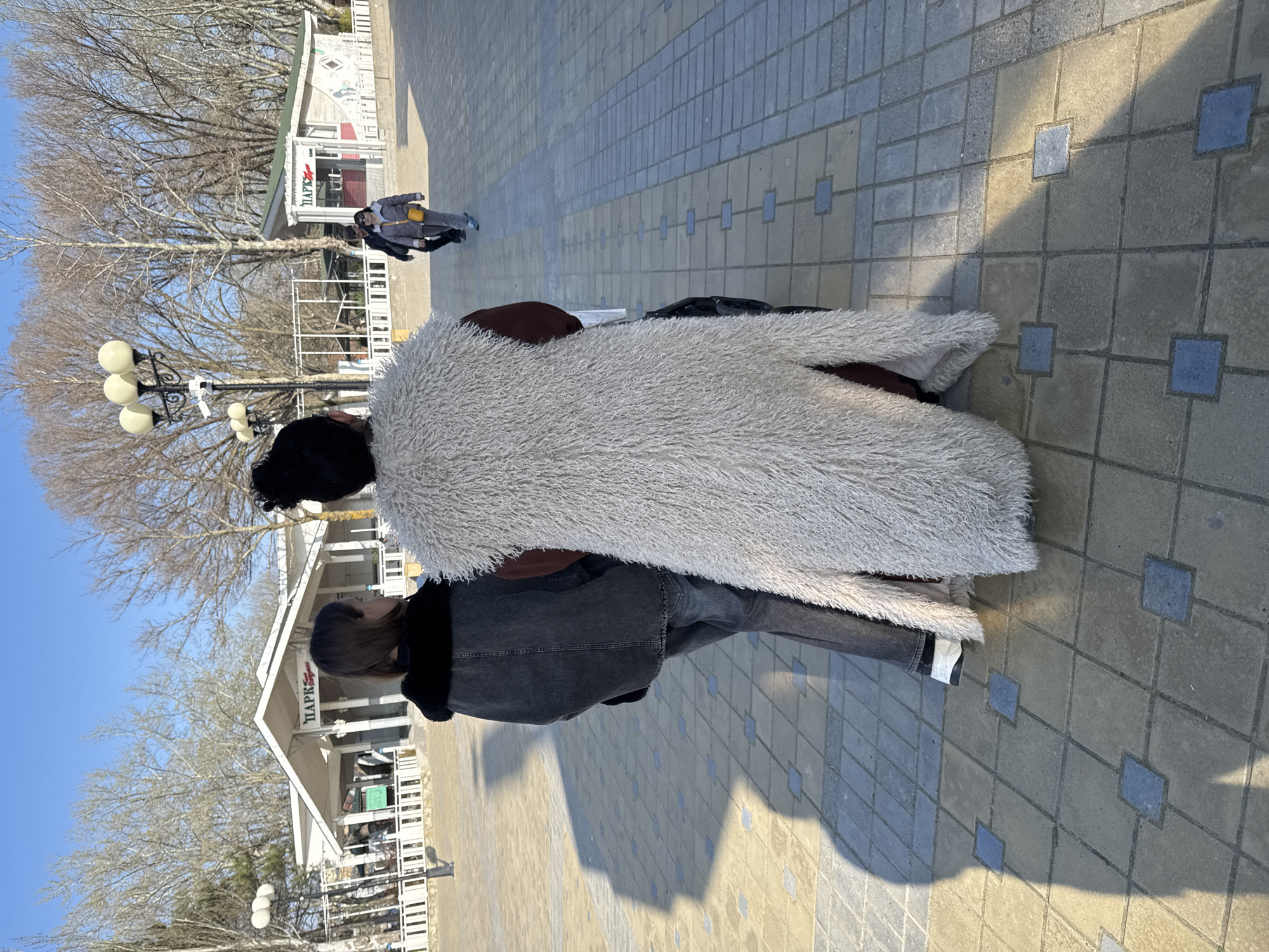 The image captures an outdoor paved area within a park in Anapa, Russia, under a clear blue sky. Two individuals are positioned in the immediate foreground, observed from behind, facing away from the camera. The person on the left wears a dark gray denim jacket with a black fur collar and dark gray trousers, displaying medium-length brown hair. The person on the right wears a long, shaggy, off-white/cream sleeveless coat over a dark reddish-brown garment, with dark hair pulled up. They stand on paving composed of light brown/tan and grey-blue interlocking square pavers. Distinct, elongated shadows are cast to the left on the paving, indicating direct sunlight from the right.
In the mid-ground, a black ornate streetlight featuring three spherical white lamps and an attached CCTV camera is visible on the left. Further right, two additional individuals walk away from the camera; one wears a dark jacket, and the other wears a light purple/gray puffer jacket and a yellow cross-body bag.
The background features two white buildings with pitched roofs, separated by white fences. The larger building on the left prominently displays "ПАРК" (PARK) signage. A smaller building with a green roof, also bearing "ПАРК" signage, is situated to the right. Numerous bare deciduous trees with visible branches dominate the background, suggesting late winter or early spring conditions.