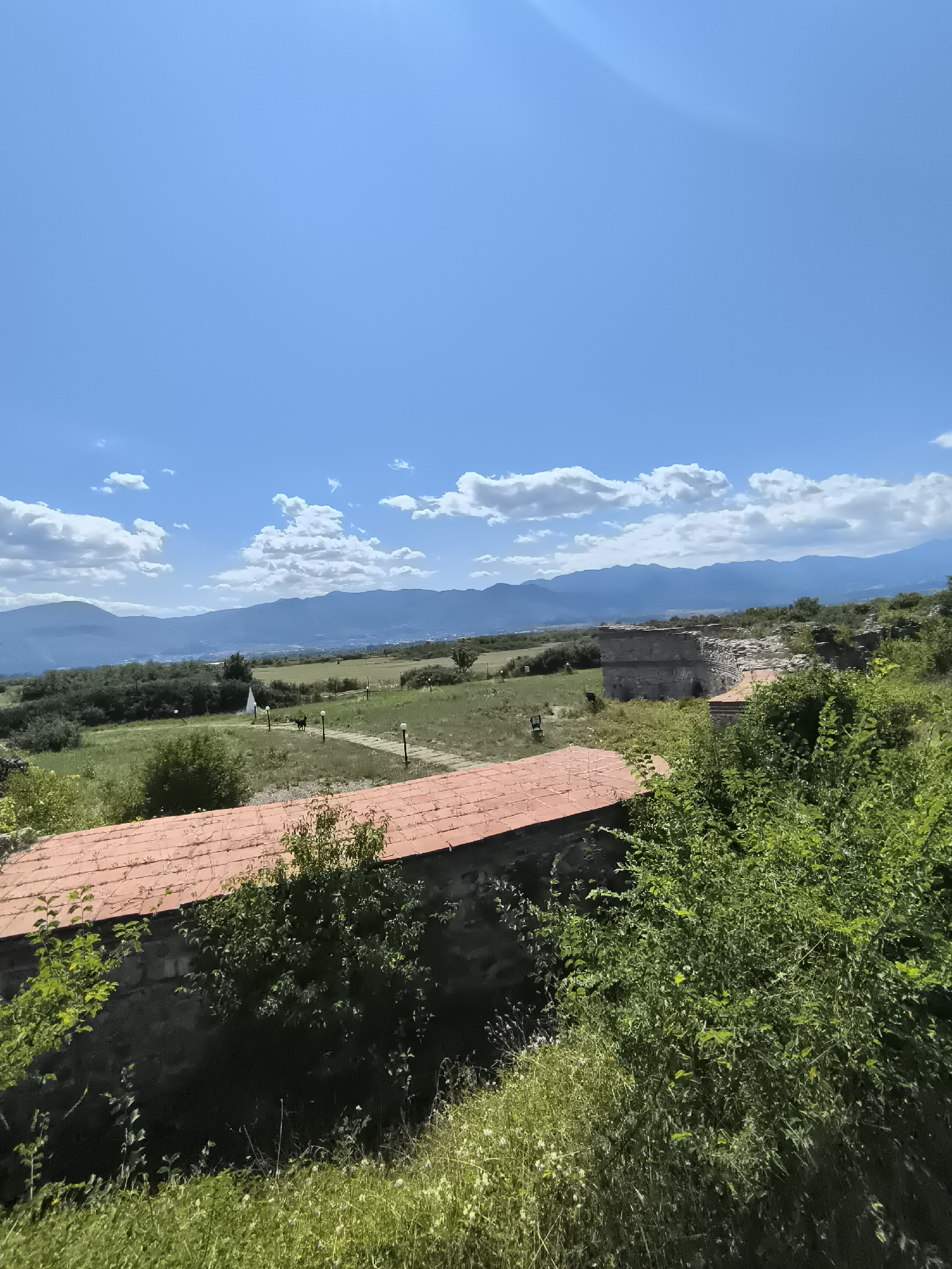 The image shows a landscape scene in Garmen, Bulgaria, under a bright blue sky with scattered white clouds. In the foreground, a red-tiled roof of a building is visible, partially obscured by lush green foliage. Behind the roof, stone ruins of a structure stand among overgrown vegetation and trees. Further in the background, a green field stretches out, dotted with sparse trees and a few lampposts. A pathway leads through the field, and a white structure or marker is present. Two dark figures, possibly animals, are situated on the pathway in the middle ground. Beyond the field, a range of blue-hued mountains forms the horizon, under the expansive sky. The overall scene depicts an outdoor, historical, and natural environment.