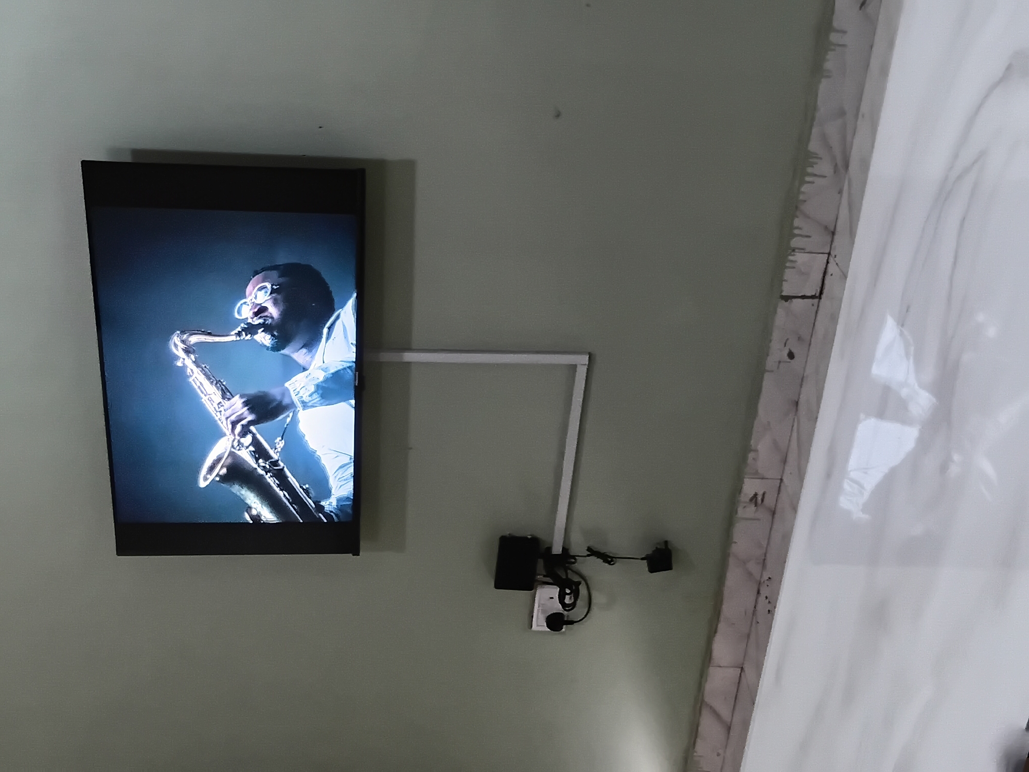 The image displays an indoor scene, likely in Barden Yana, Nigeria. A flat-screen television is mounted on a light green wall, showing a dark-skinned man wearing glasses and a light-colored shirt, intently playing a saxophone. The man on the screen is illuminated, highlighting his focus on the instrument.
To the right of the television, a white, L-shaped wall-mounted light fixture extends. Below this fixture, several electronic devices are installed on the light green wall: a small black box, a white power outlet or switch, and a black power adapter with connected cables, indicating a functional setup. To the far right of the image, a vertical white surface, possibly a tiled wall or column, is visible. This surface has distinct dark lines, likely grout or structural imperfections, creating a textural contrast with the smooth green wall. No specific text is visible on any objects within the frame. The overall scene suggests a contemporary interior space, possibly a living room or common area.