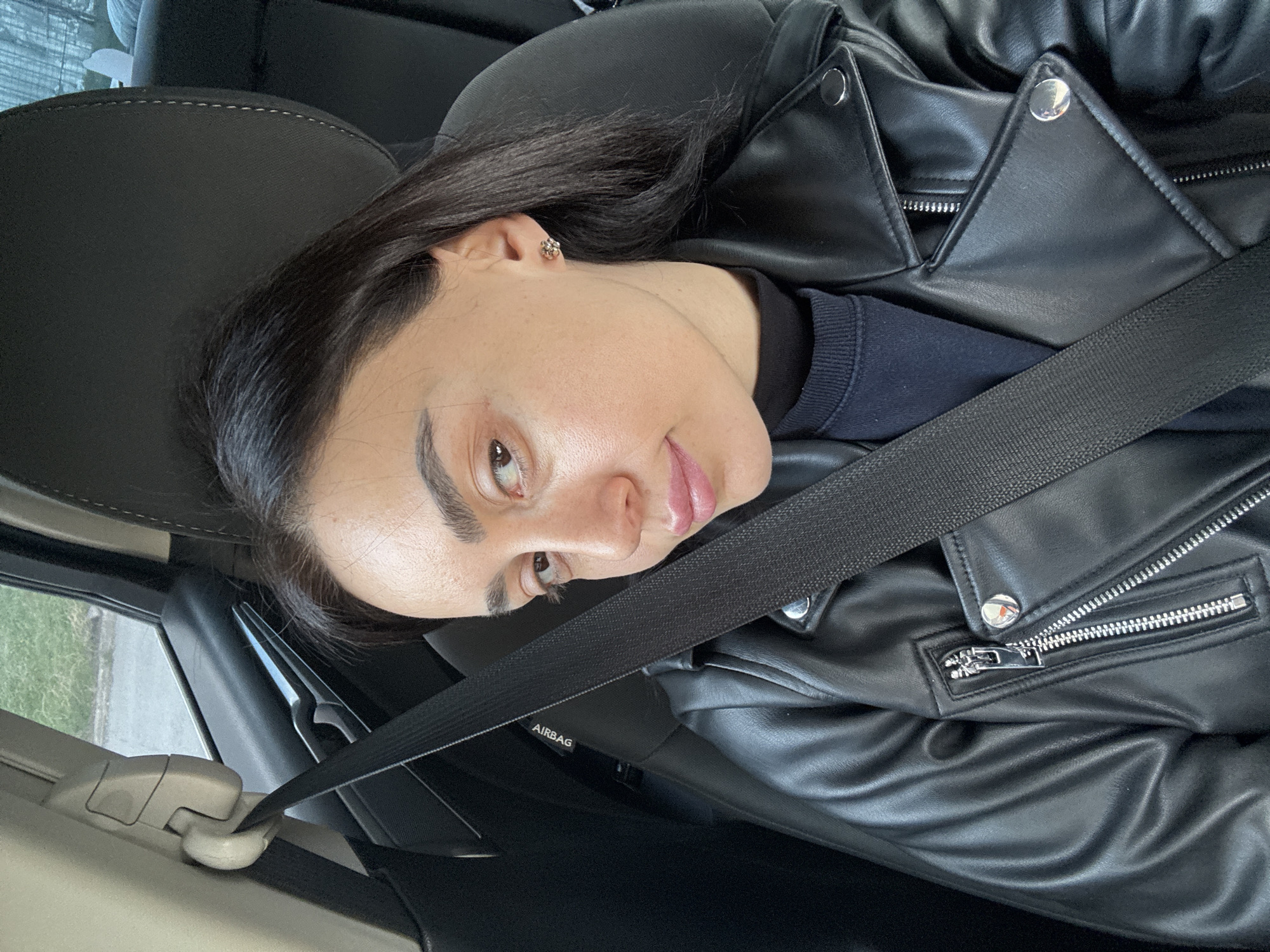 A woman is sitting in a car, wearing a black leather jacket and a black top. She has dark hair and is wearing a seatbelt. The car appears to be in motion, but the location in the city of Tbilisi, Georgia, cannot be determined.
