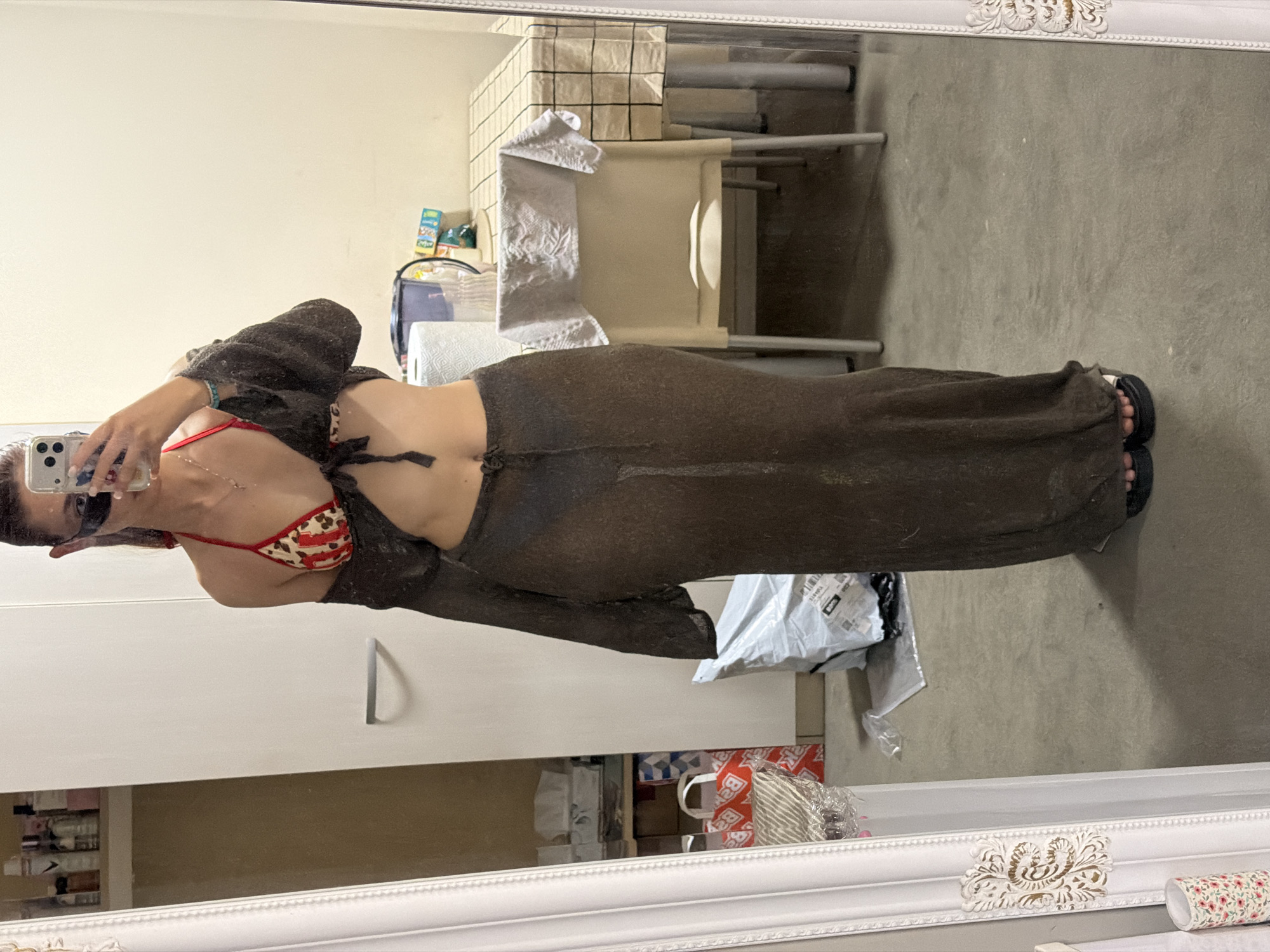 A mirrored self-portrait of a woman wearing a bikini top with a red and white pattern, a long brown sheer dress and a matching cardigan is captured. She holds a smartphone up to take the picture. The room behind includes a cabinet and a chair with a checkered cover. Packages and other items are visible in the background. The location is a room with furniture, possibly an apartment in Sofia, Bulgaria, though the environment is not detailed enough for precise geocoding.