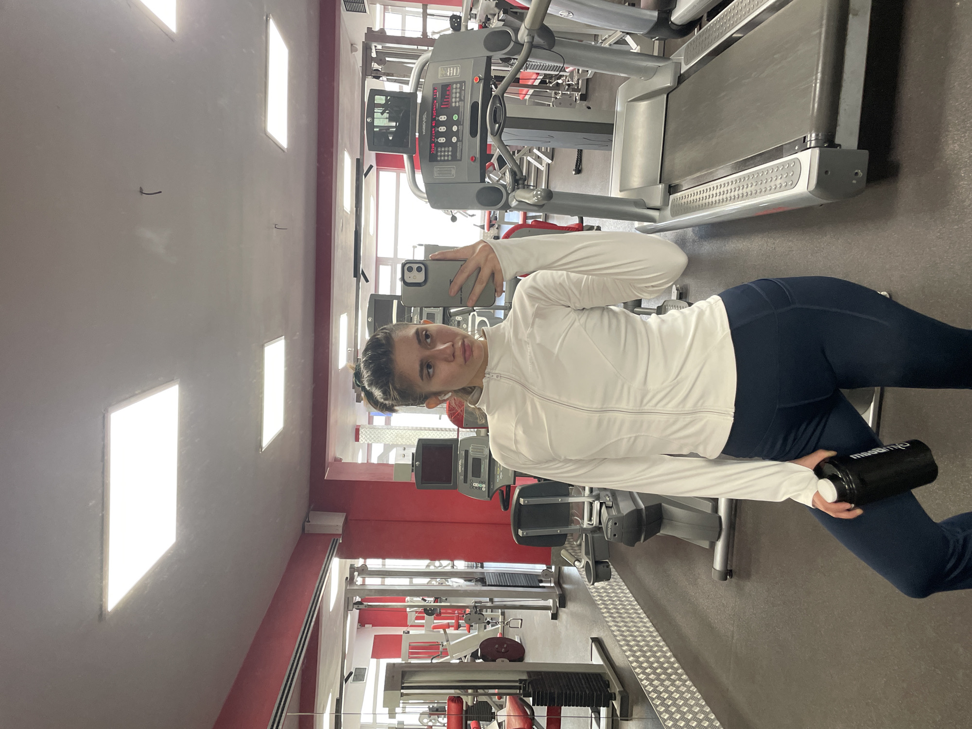 The image is a selfie taken in a gym, likely located in Sofia, Bulgaria, given the red and white color scheme common in Bulgarian athletic facilities. The subject is a young woman wearing a white long-sleeved jacket and dark blue leggings. She is holding a black water bottle and a smartphone, presumably to take the selfie. Behind her is a treadmill, other gym equipment, and the interior of a fitness center with multiple exercise machines arranged in rows. Natural light enters the space through high windows.