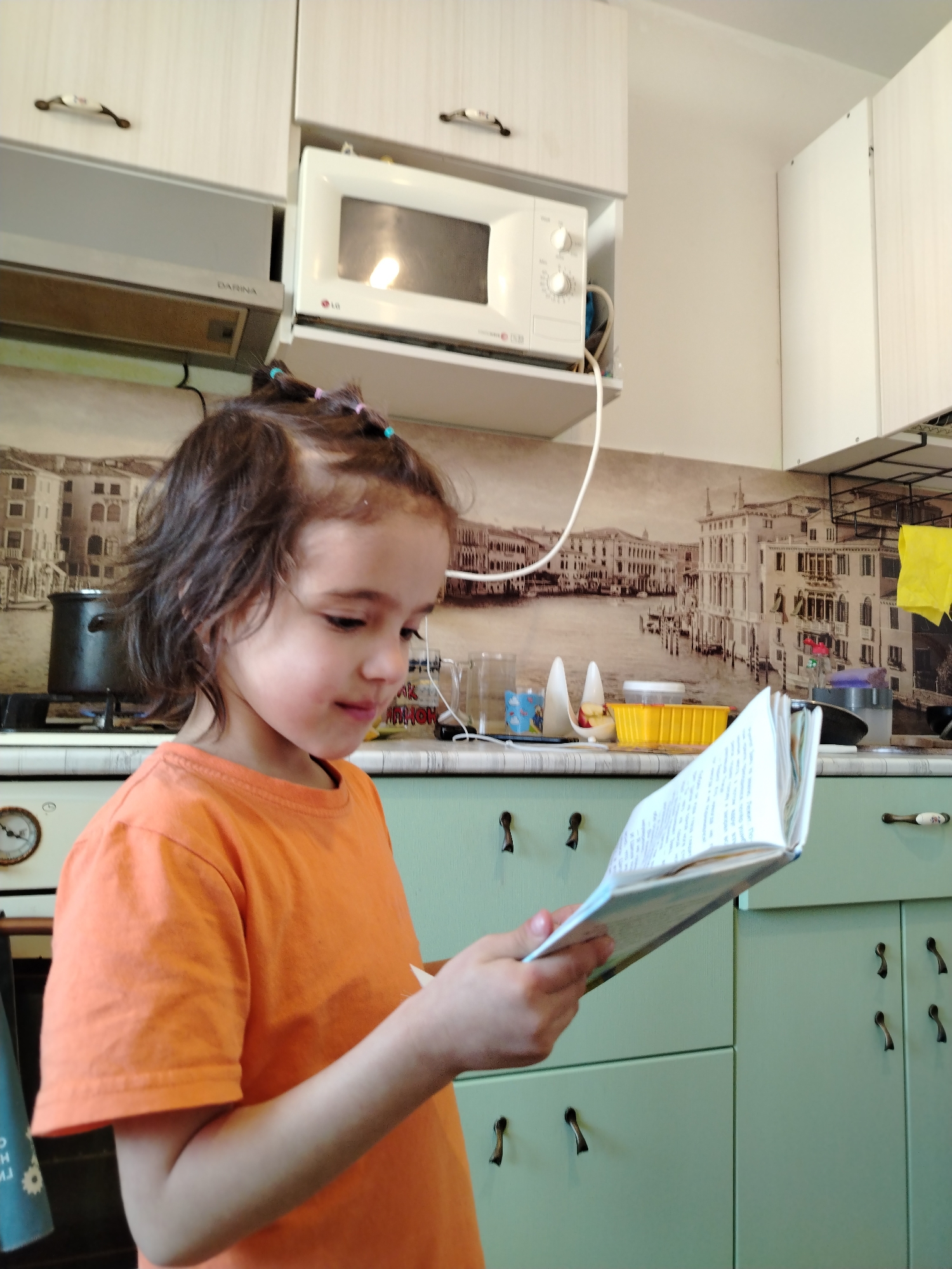 A young child, likely a girl, is captured in a home kitchen setting, engrossed in reading an open book. She has dark brown hair, styled with small pigtails adorned with colorful elastic bands, and is wearing a vibrant orange short-sleeved t-shirt. Her head is tilted downwards, and her expression indicates focus and concentration on the book's pages.
The kitchen features light green lower cabinets and white upper cabinets, both equipped with distinctive dark, curved handles. A light-colored countertop separates the upper and lower cabinetry. The wall above the countertop is decorated with a sepia-toned wallpaper or backsplash depicting a scenic view of Venice, showing canals and architectural buildings.
On the countertop, various kitchen items are visible, including several clear glass jars, a yellow plastic container, and a white power cord. A black pot is placed on a stove burner in the background. Above the stove, a silver range hood with the brand "DARINA" is installed. To the right of the range hood, a white LG microwave oven is mounted on a shelf. A yellow cloth hangs on a rack to the right, and a round, clock-like object is partially visible on the far left. The scene portrays a quiet, daytime activity in a functional home kitchen.